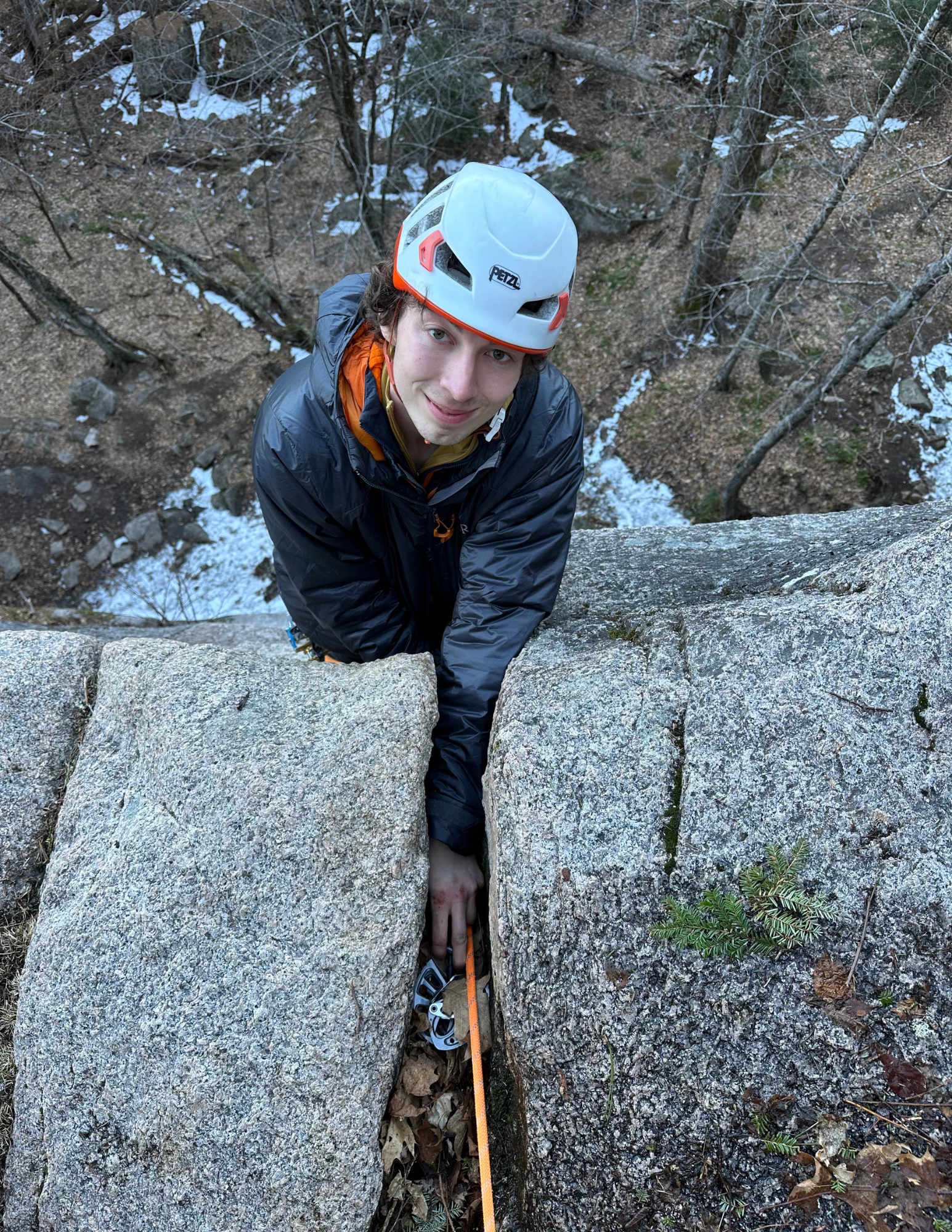 A man in a white climbing helmet and black jacket climbs a granite rock face, holding onto a crack with climbing gear attached to his harness.