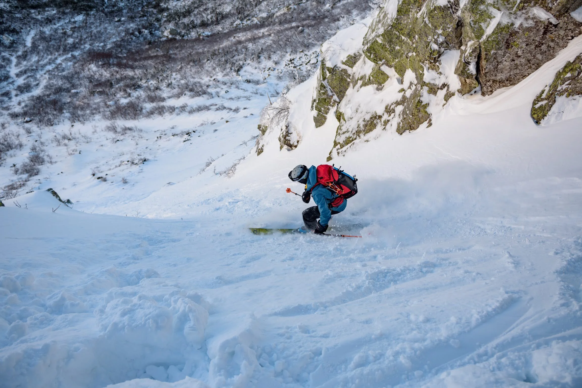 A person wearing a blue jacket and black pants snowboarding down a snowy mountain slope with a rocky cliff in the background.