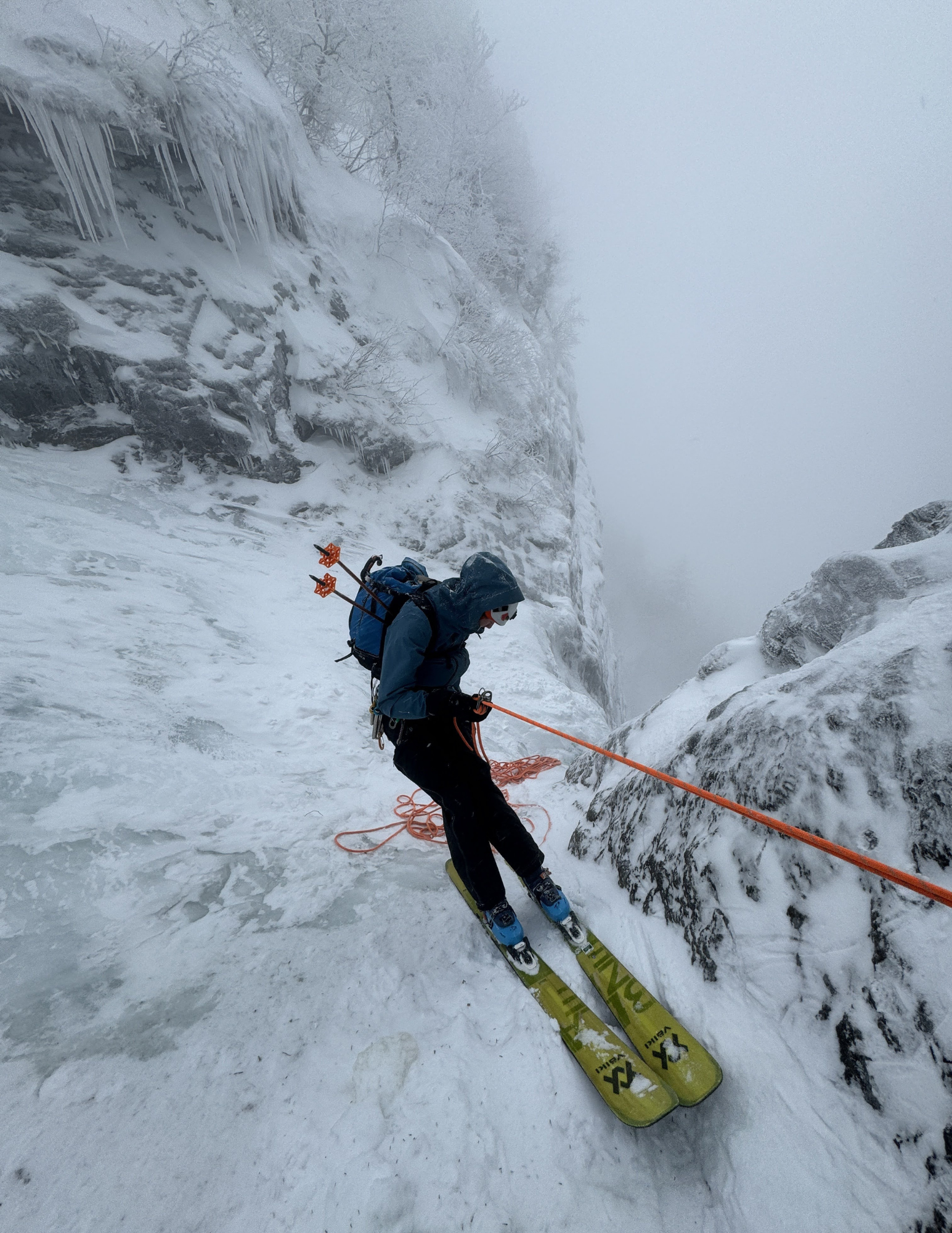 A person in winter gear skiing down a snowy, icy slope with a backpack and an orange safety rope, surrounded by snow-covered rocks and trees, in a foggy winter landscape.