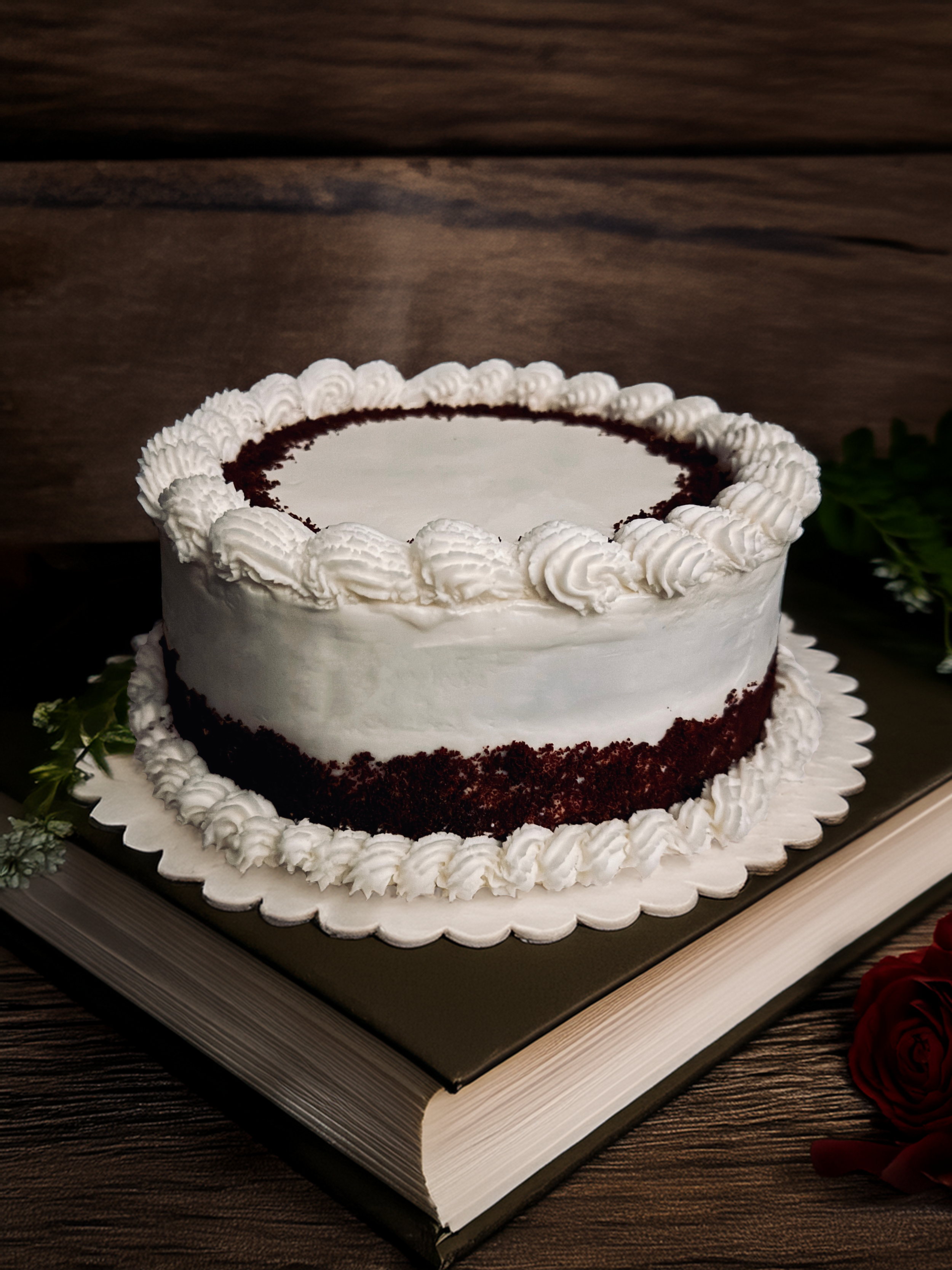 A round layered cake with white frosting and decorative piped frosting border, sitting on a silver tray with a book underneath. Red roses are visible on the side and background.