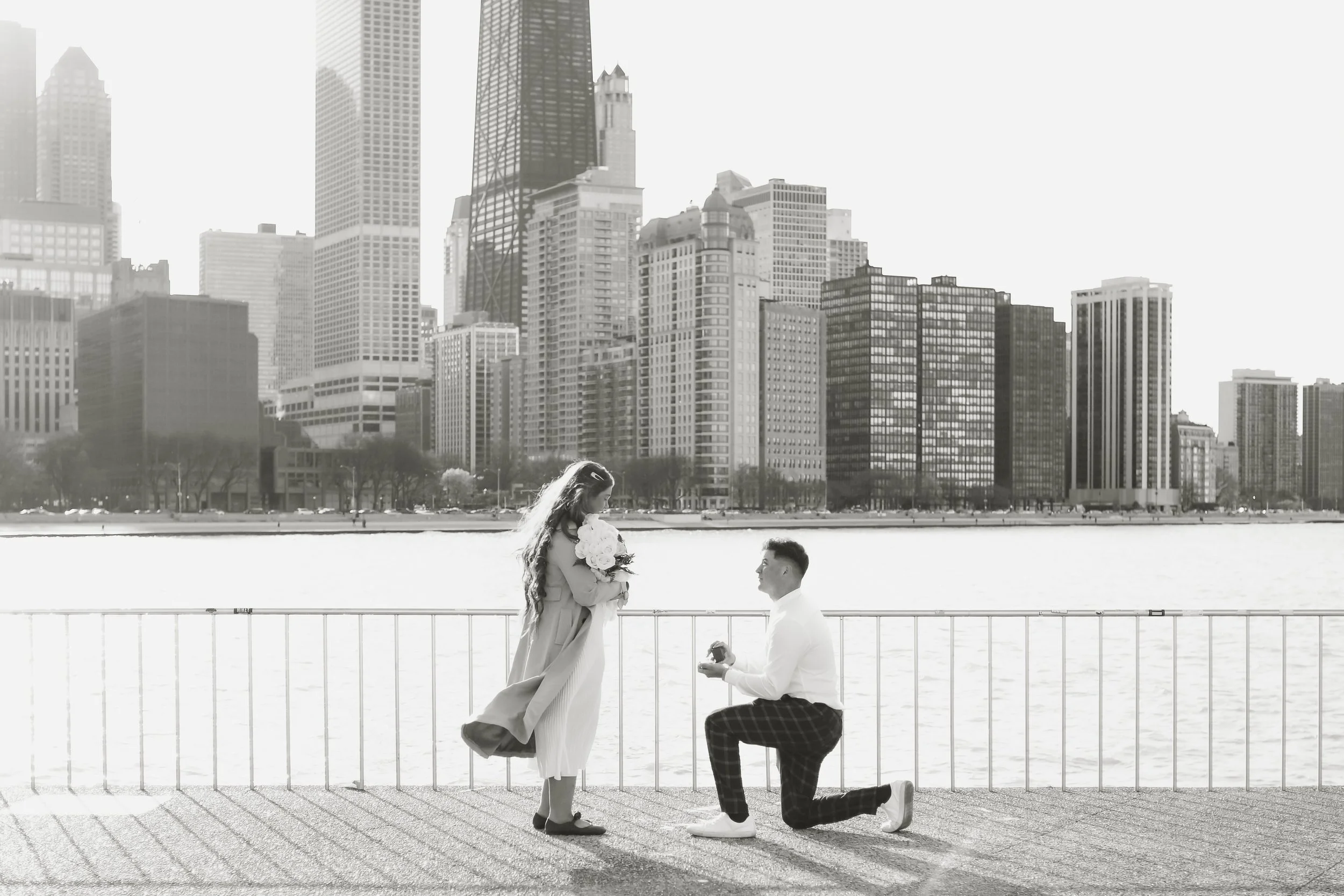 A man proposing to a woman by a waterfront with a Chicago city skyline in the background, in black and white.