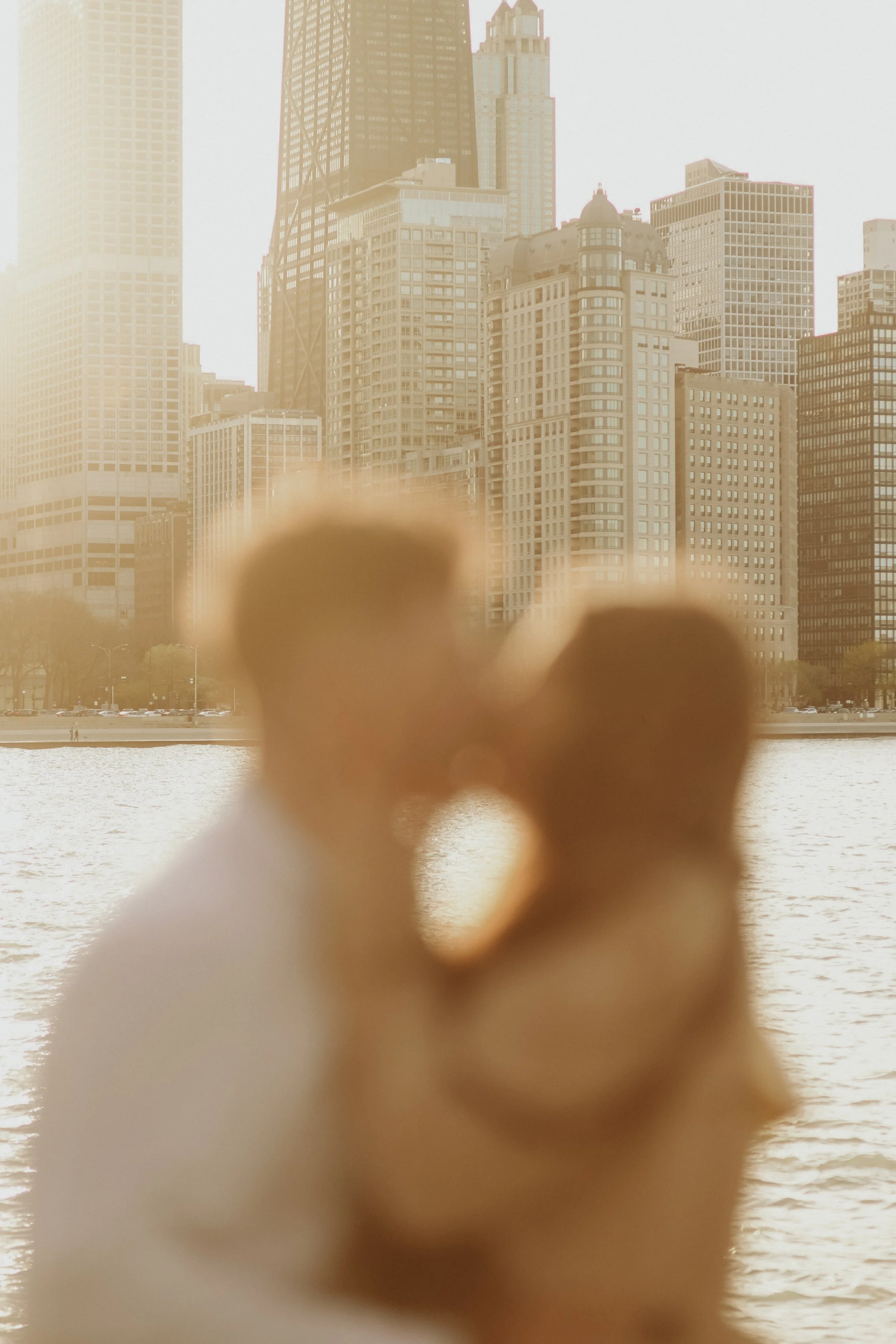 A couple holding hands, out of focus, with a city skyline and a body of water in the background.