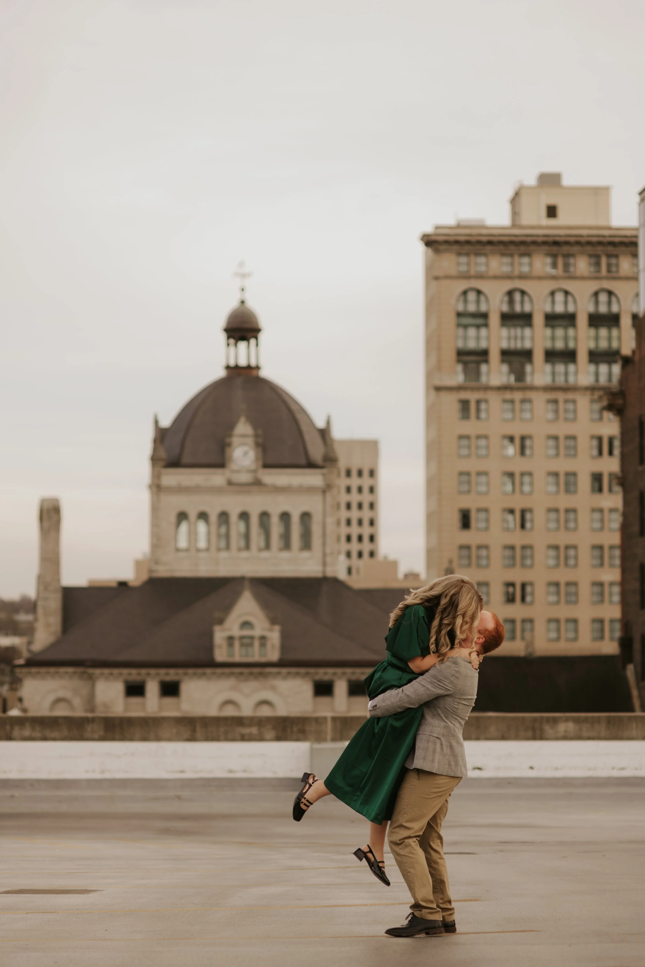 A man lifts a woman in a green dress on a rooftop in front of city buildings, sharing a kiss.