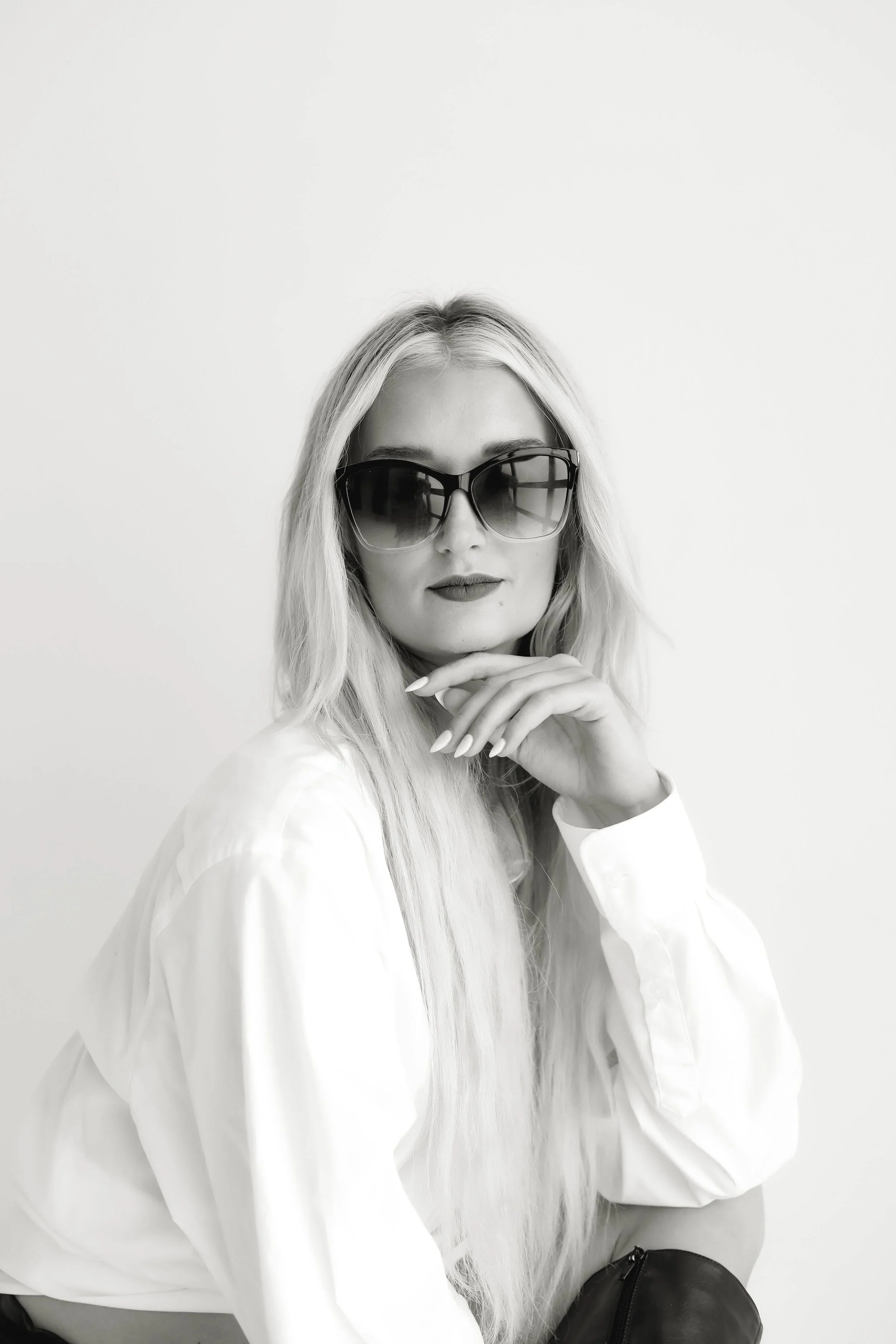 Black and white photo of a woman with long blonde hair wearing sunglasses, a white shirt, and lipstick, sitting against a plain white background.