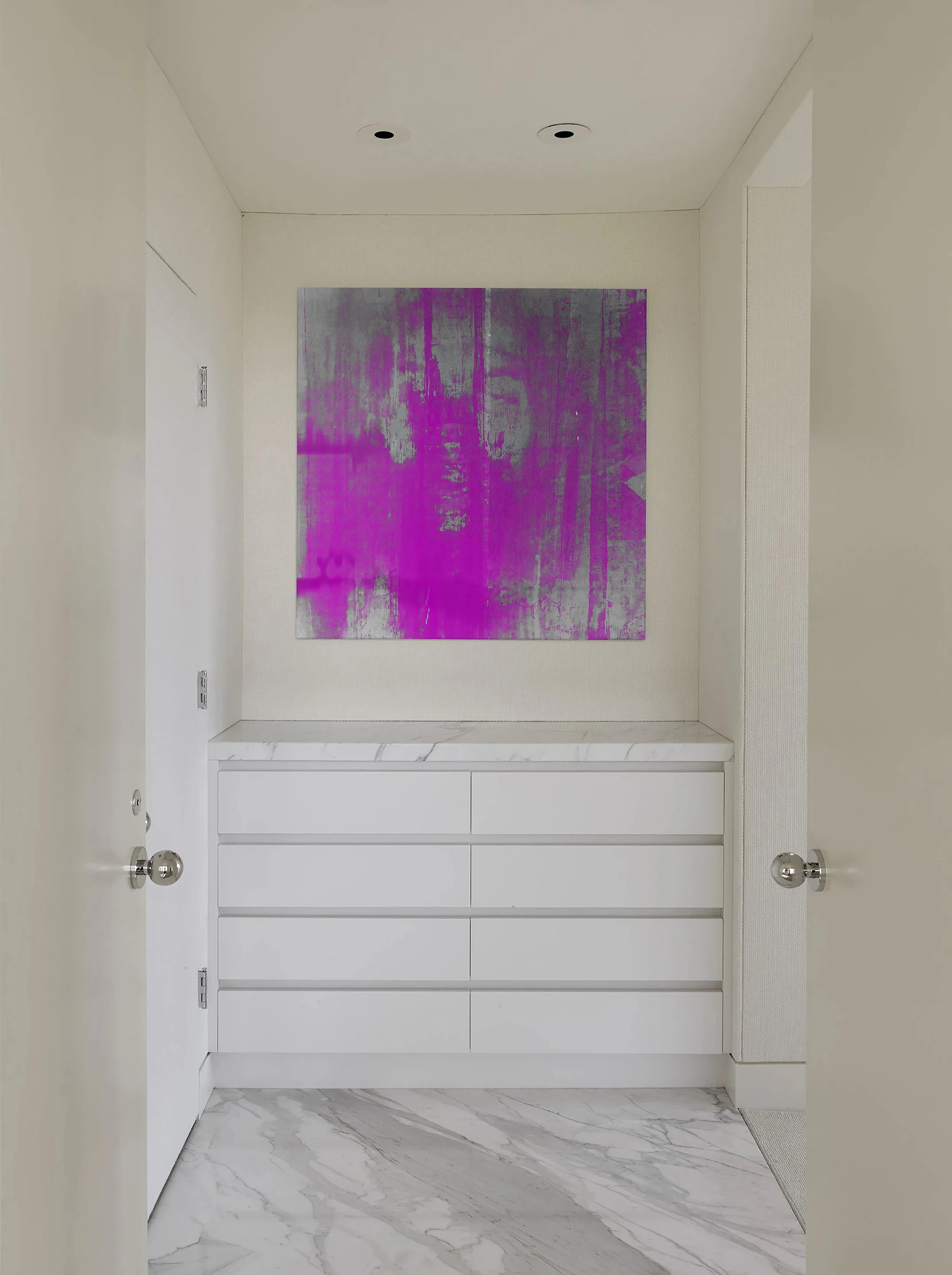 A modern entryway with white walls, white marble flooring, a white dresser with six drawers, and a square abstract artwork with purple and gray tones hanging on the wall.