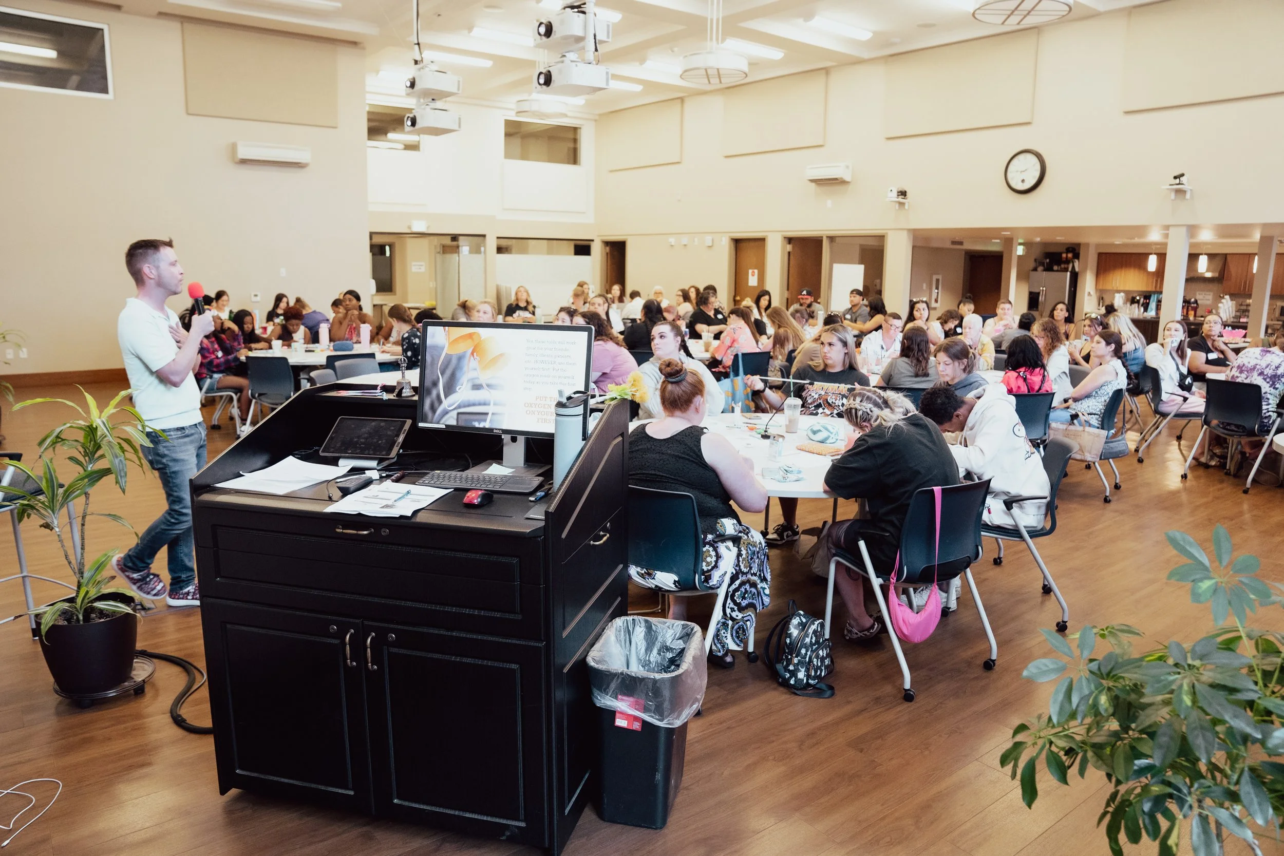 A speaker stands in front of a large audience in a spacious conference room, holding a microphone. The audience is seated at round tables, listening attentively. The room has high ceilings, with projectors hanging from above and a clock on the wall.