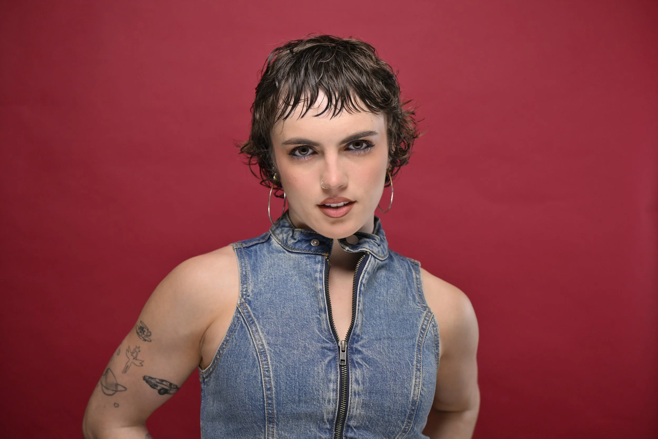 Person with short, wet-look hair, wearing a sleeveless denim top and hoop earrings, against a red background.