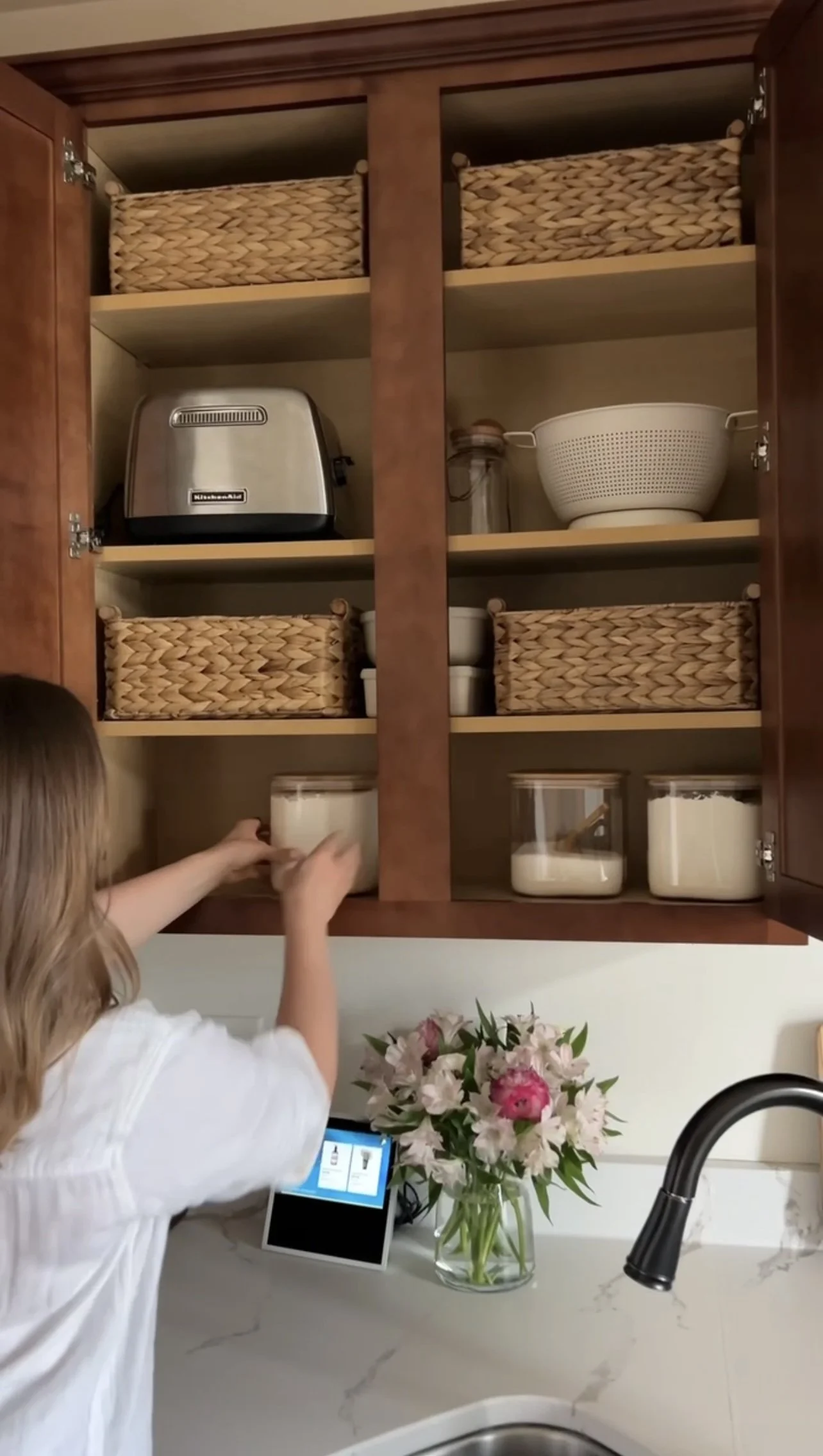 Open organized kitchen cabinet with baskets, a toaster, a colander, glass jars, and containers of ingredients, with a woman reaching into a container on the kitchen counter.