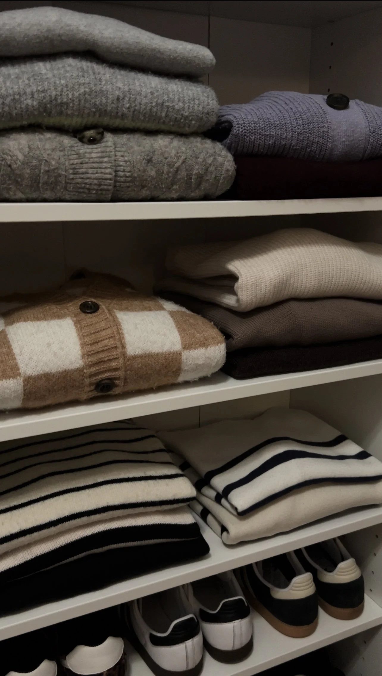 Organization of folded sweaters, socks, and sneakers on white shelves in a closet.