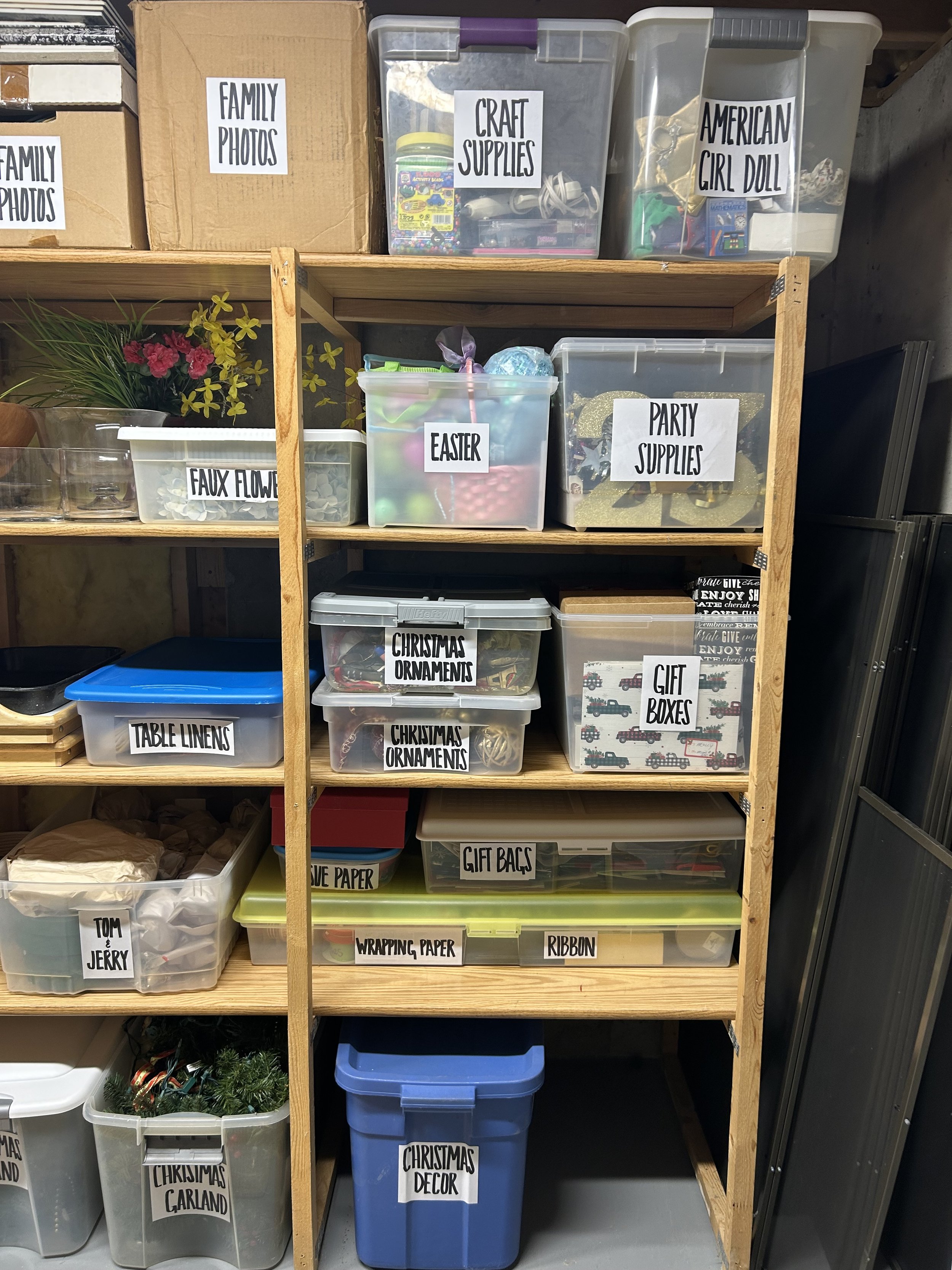 Wooden shelving unit with labeled plastic storage bins containing family photos, craft supplies, items for an American girl doll, faux flowers, Easter decorations, party supplies, Christmas ornaments, gift boxes, gift bags, wrapping paper, ribbon, Ch