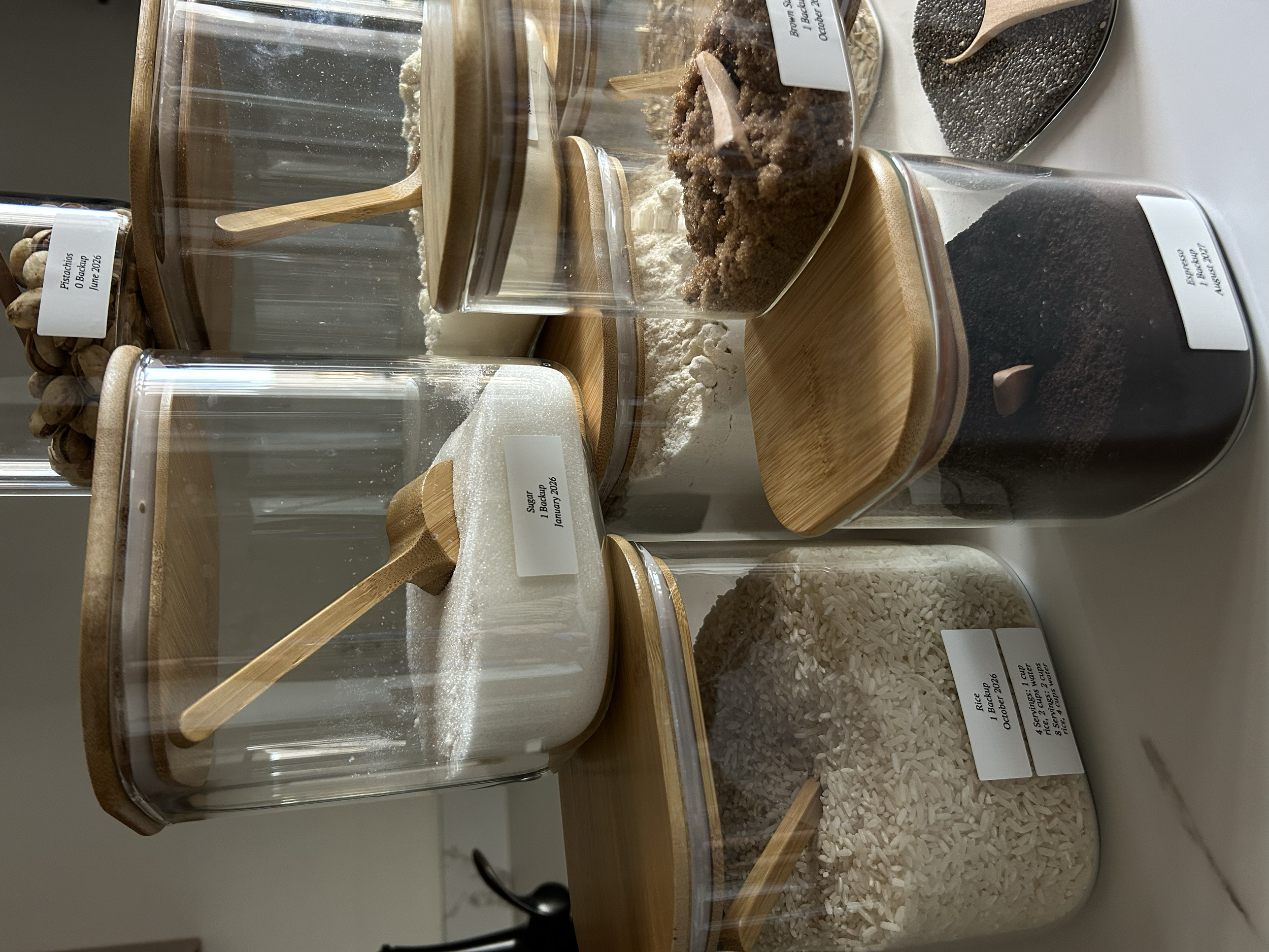 Organized  various dry food ingredients stored in glass jars with wooden lids, including sugar, rice, cocoa powder, and brownie ingredients, with small wooden spoons inside each jar.