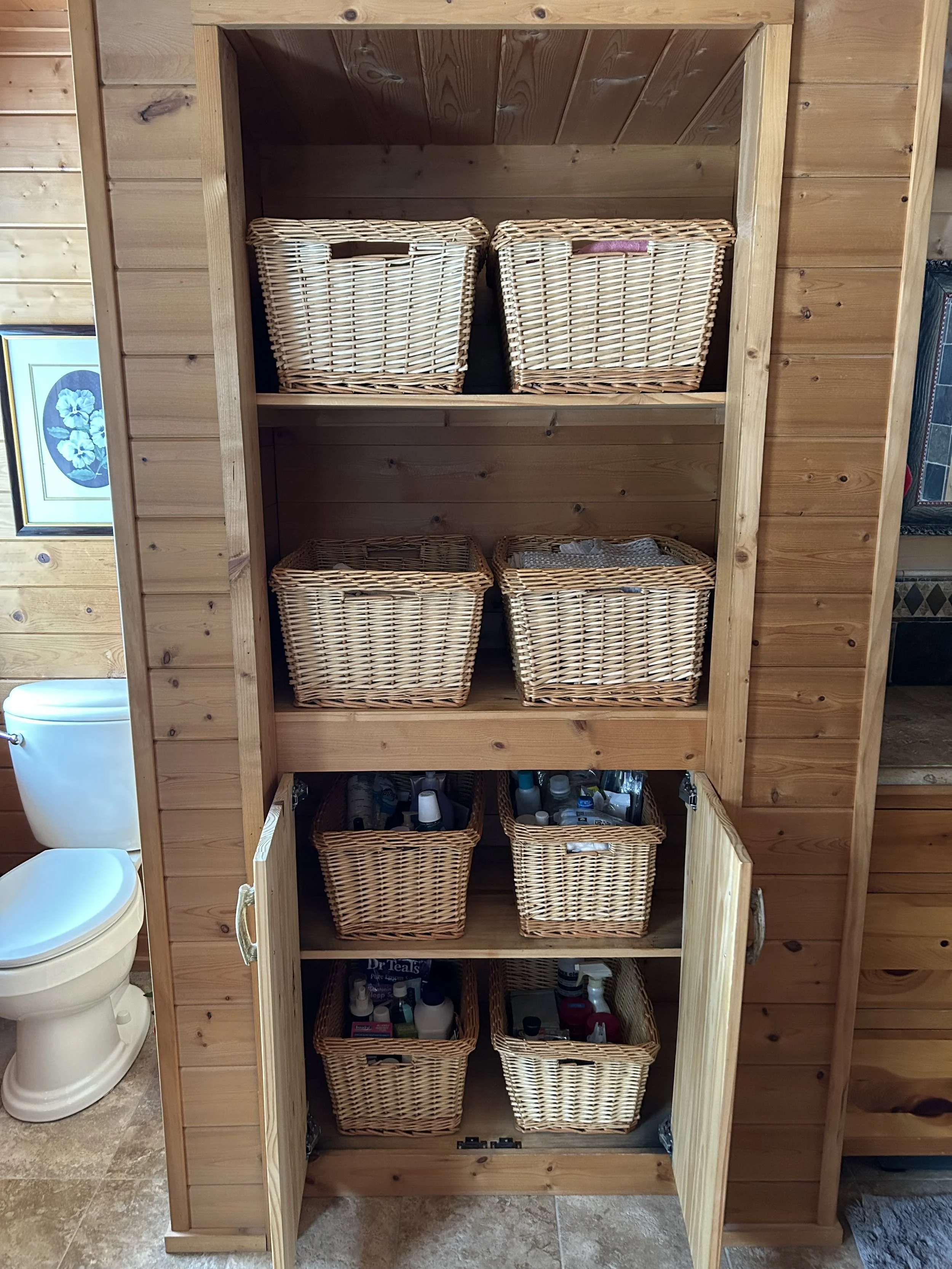 A wooden cabinet with six woven baskets filled with toiletries and supplies in a bathroom, with a toilet visible to the left.