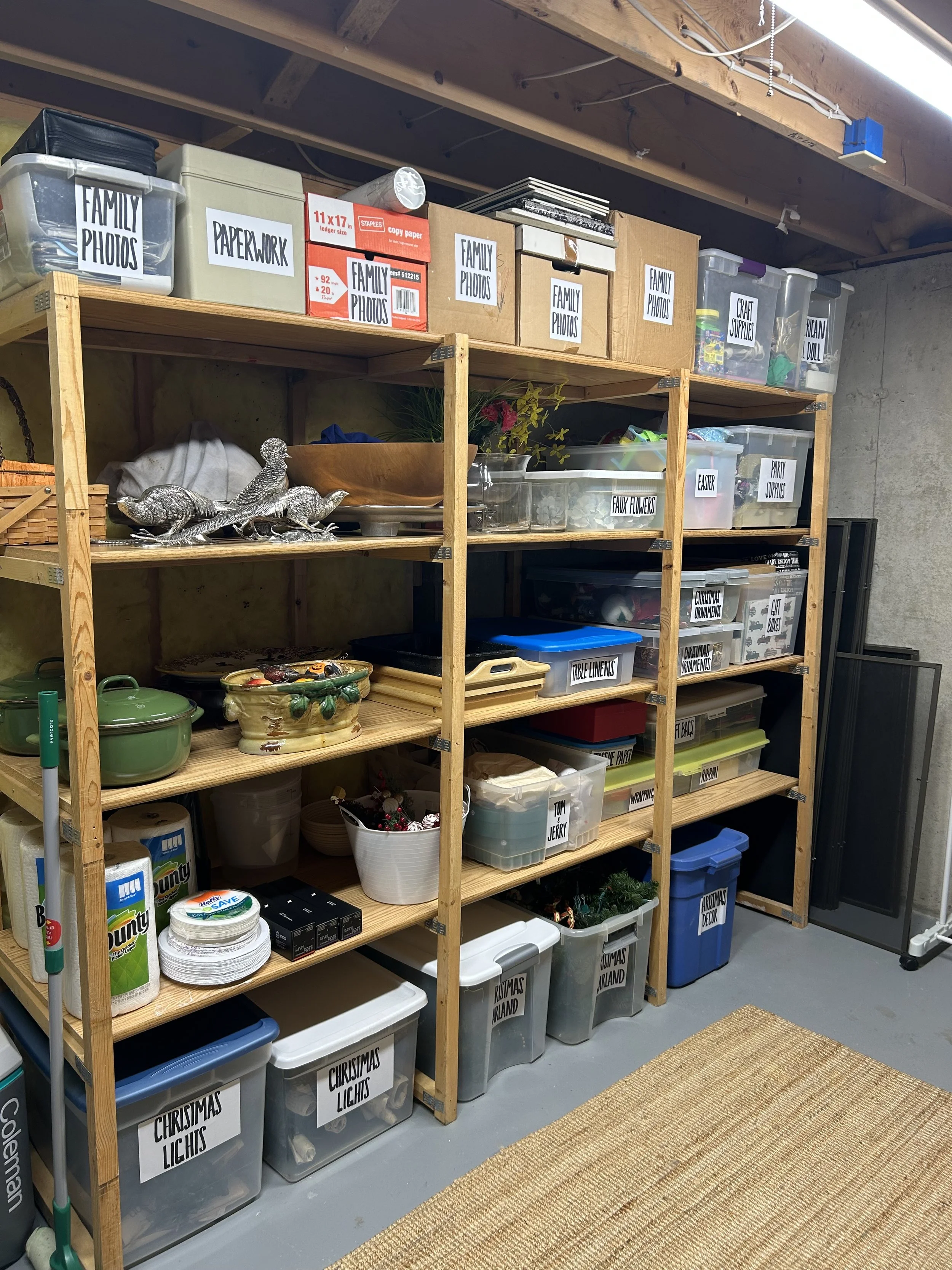 A storage shelving unit filled with labeled plastic bins, boxes, and decorative items, including family photos, craft supplies, faux flowers, and holiday decorations, in a basement or garage.