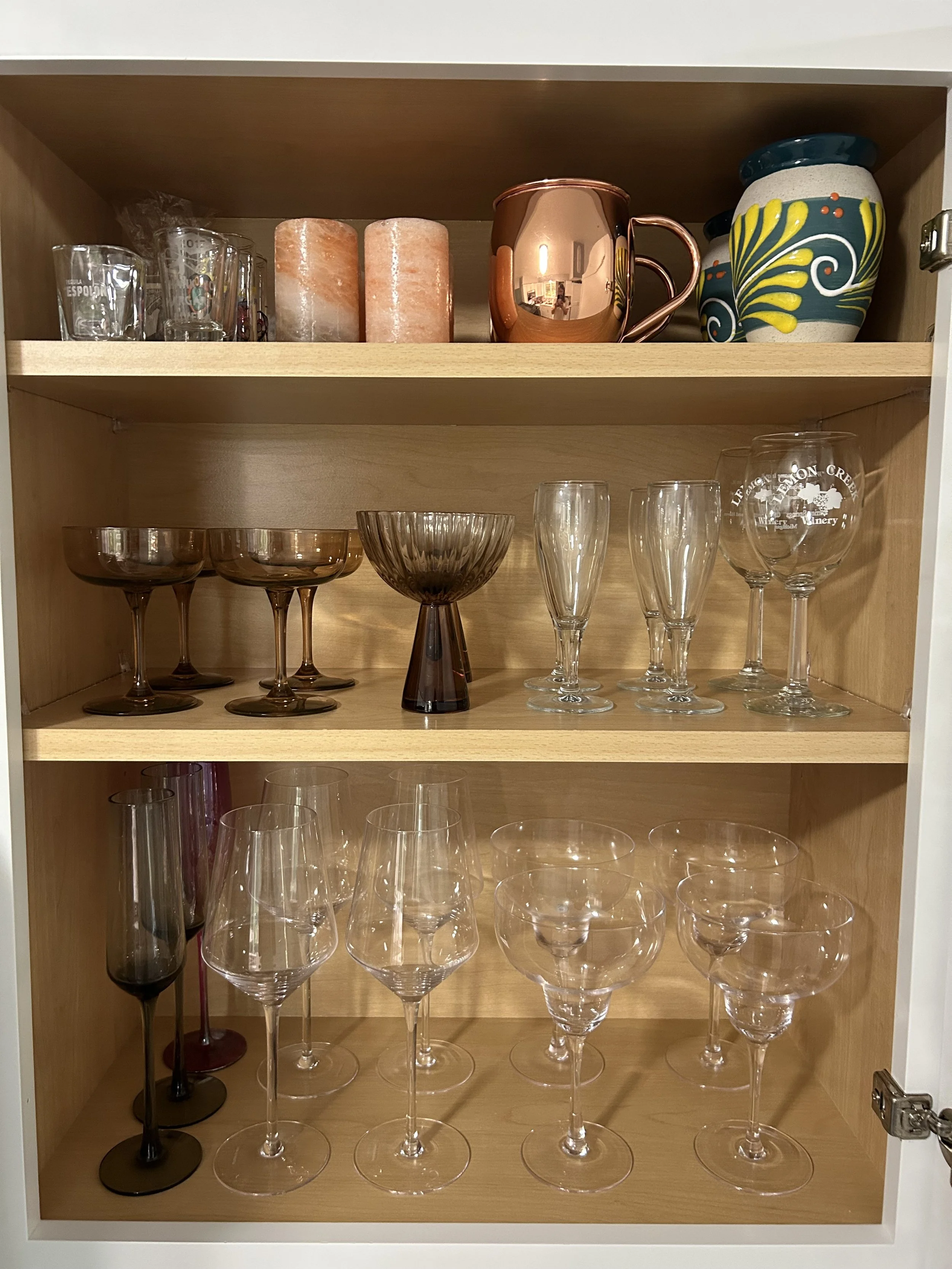 An organized wooden cabinet with three shelves filled with various glassware and decorative items, including wine glasses, champagne flutes, and a copper mug.