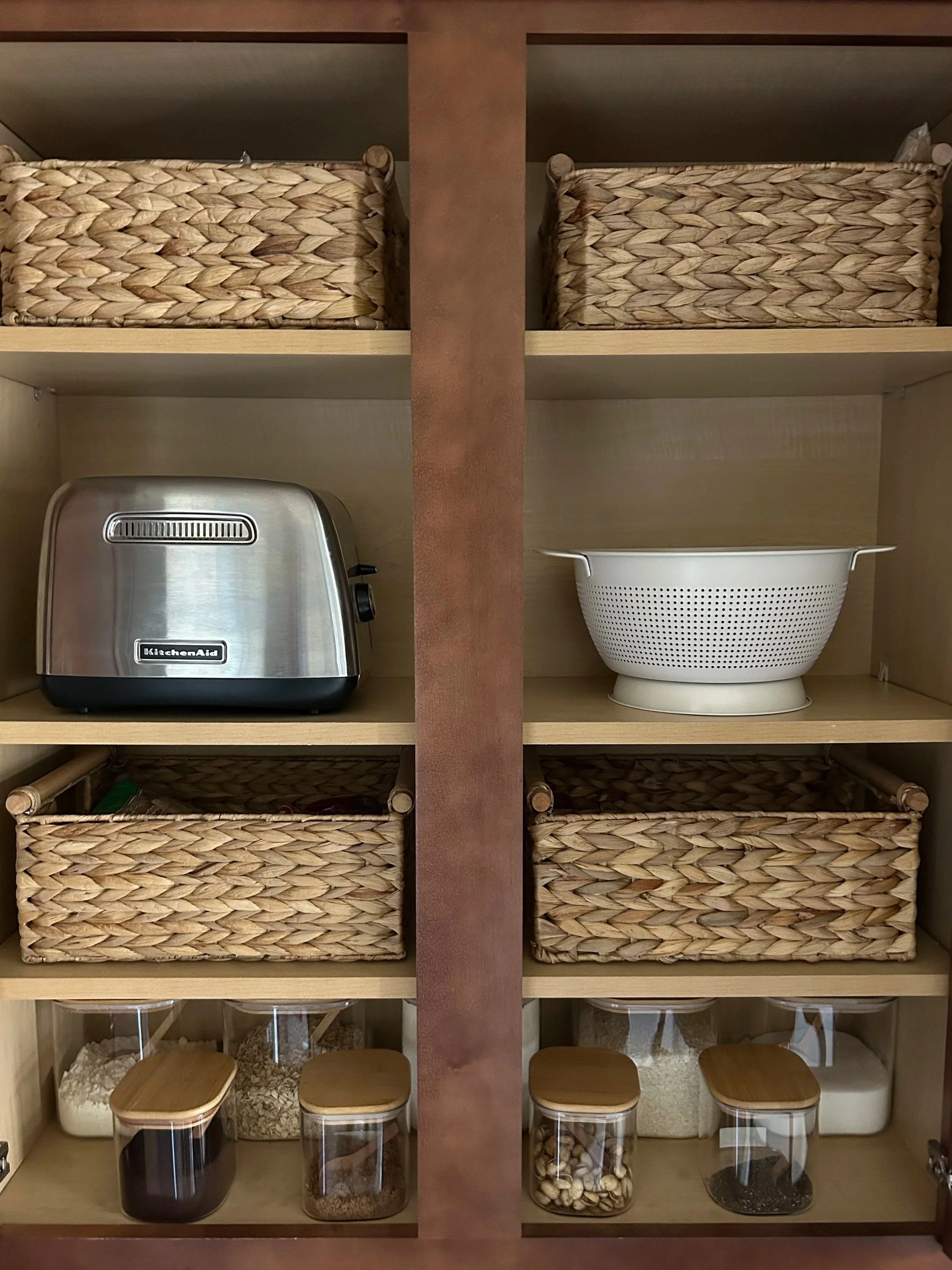 Organized kitchen pantry with woven baskets, a stainless steel toaster, a white colander, and glass jars filled with various ingredients.
