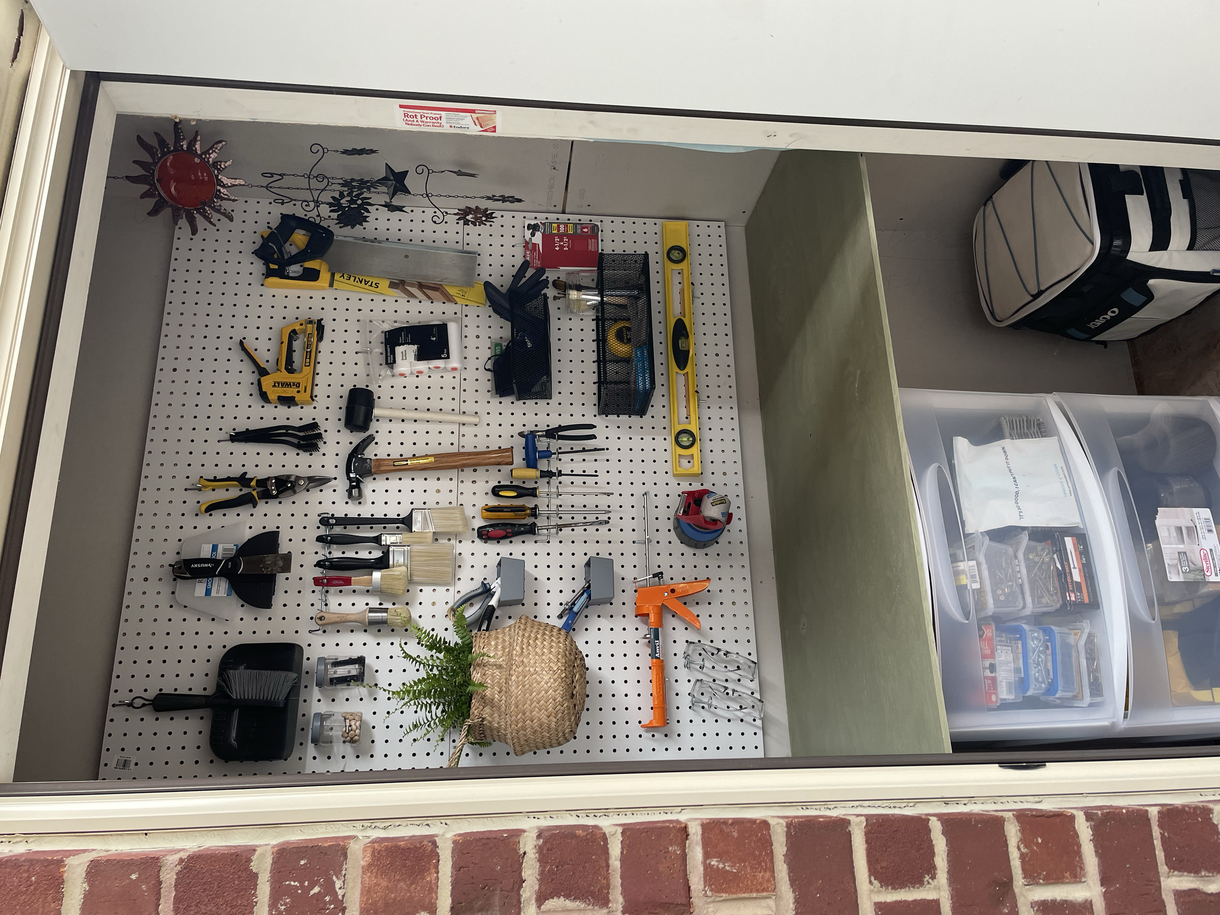 A small organized garage with a pegboard wall displaying various tools including a level, a saw, and screwdrivers. There is a plant in a woven basket, a level, a pipe clamp, and other small tools on the pegboard. The garage has a brick wall at the bo