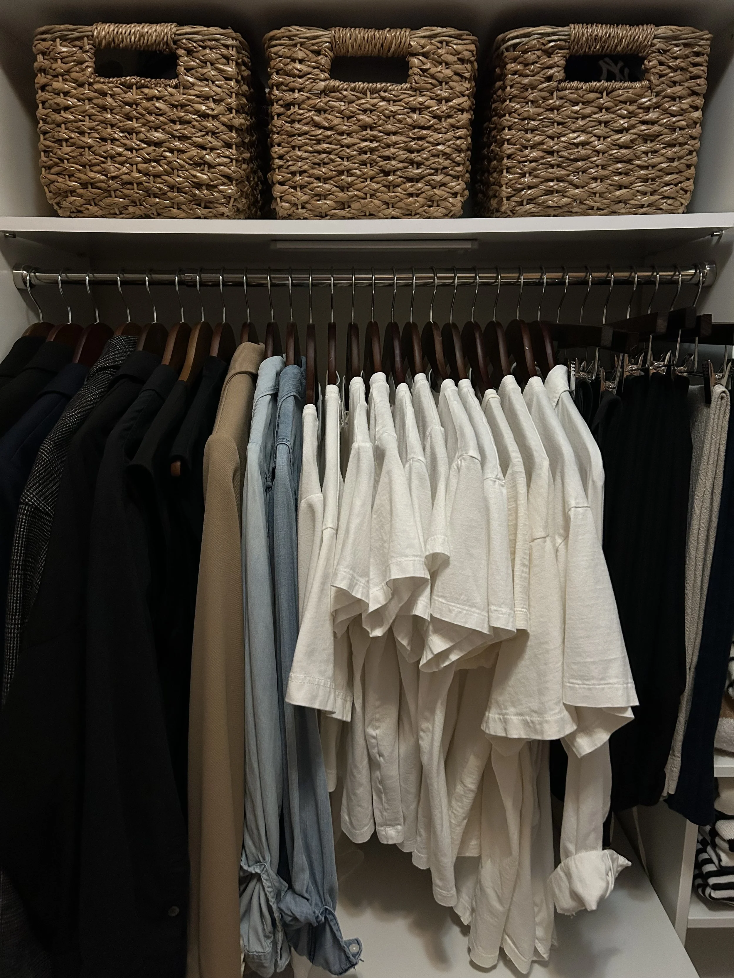 Organized closet with clothing on hangers including black, beige, light blue, white, and black pants, with woven baskets on the top shelf.