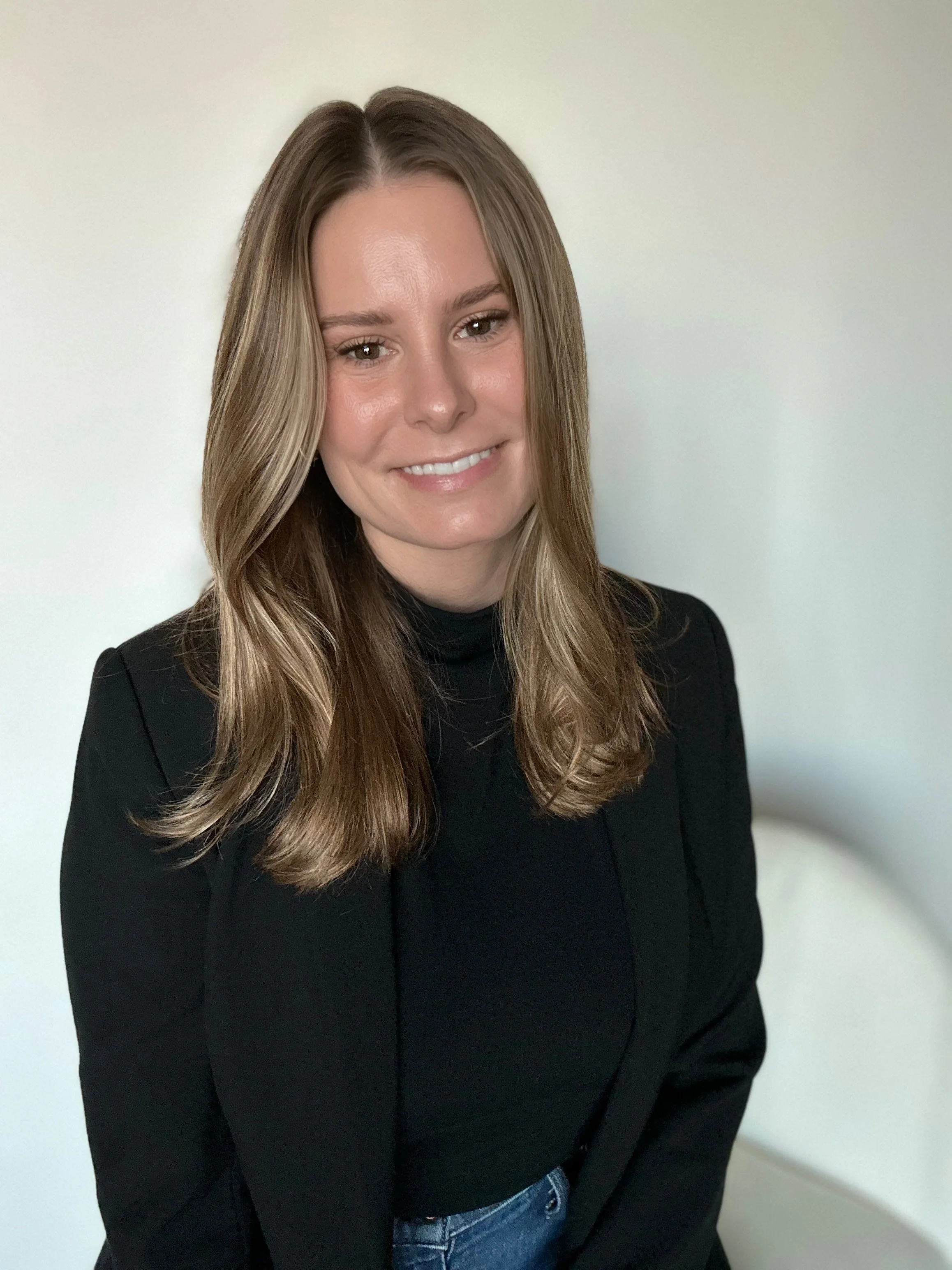 A woman with long, wavy light brown hair and fair skin, smiling, wearing a black turtleneck and blazer, against a neutral background.