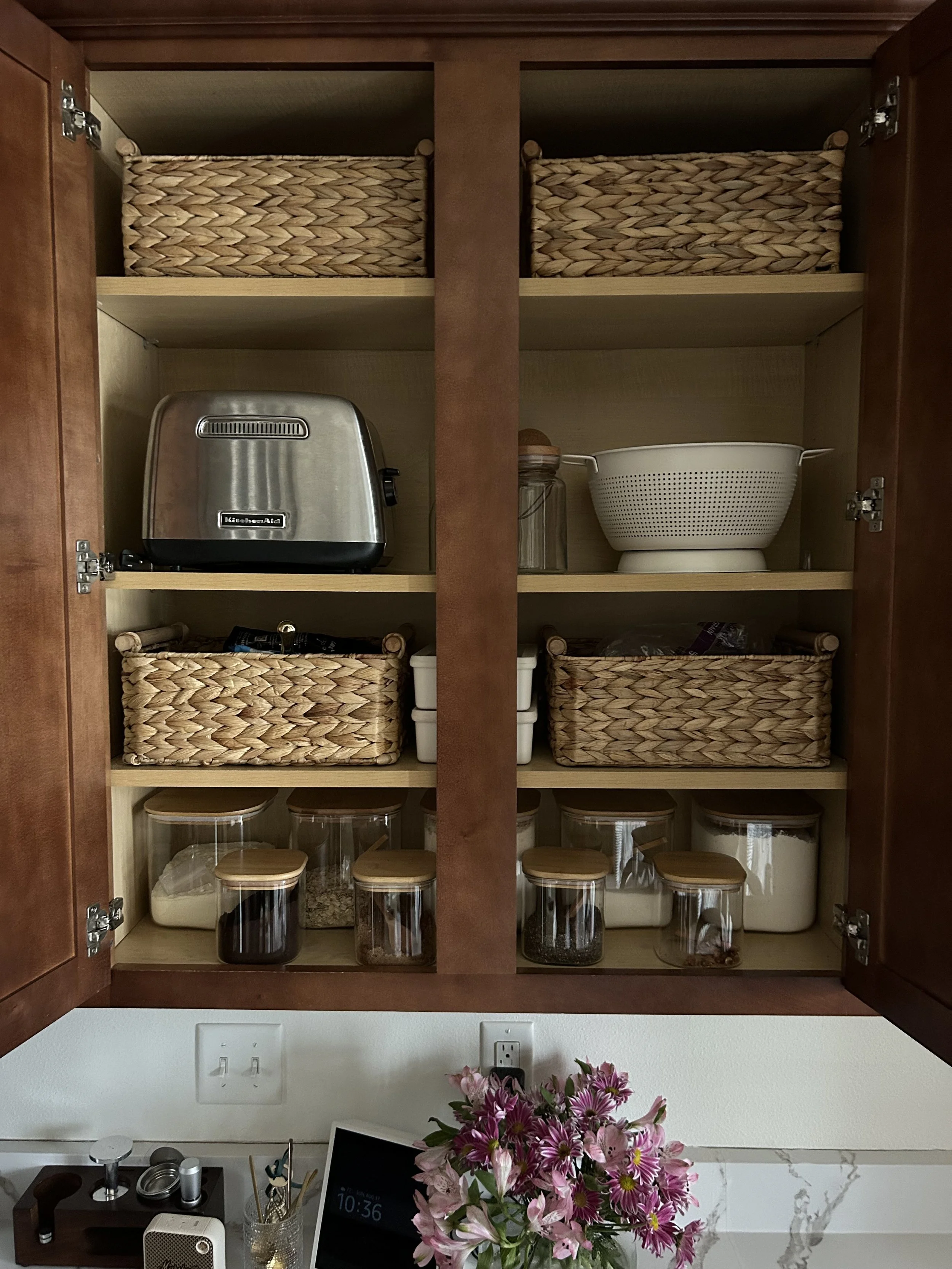 Open organized kitchen cabinet with woven baskets, a toaster, a colander, glass jars with food, and a pink flower arrangement on the countertop below.