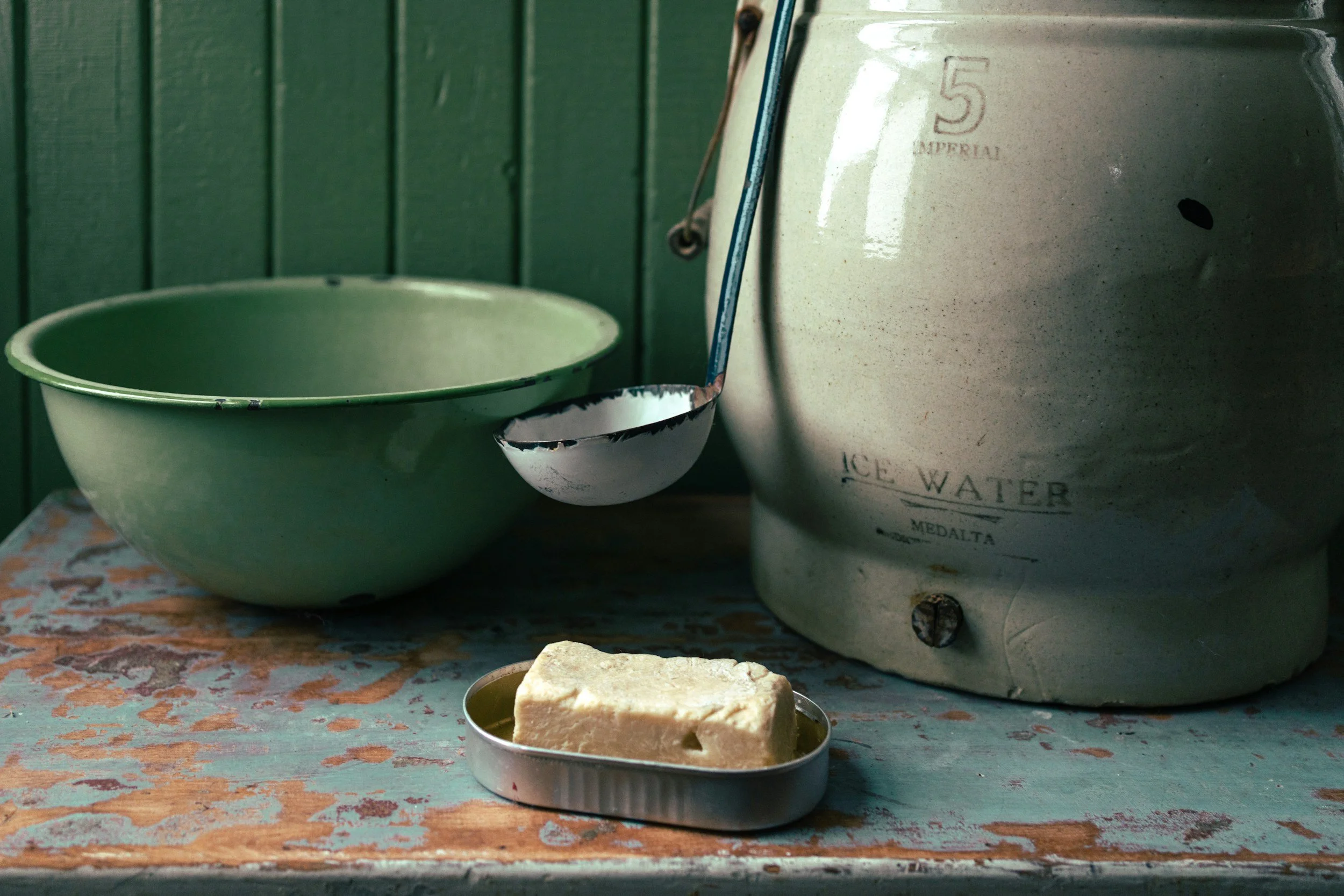 A vintage kitchen scene with a green enamel bowl, a ladle, a can of sardines, and an old ice water dispenser on a worn wooden table against a green-paneled wall.