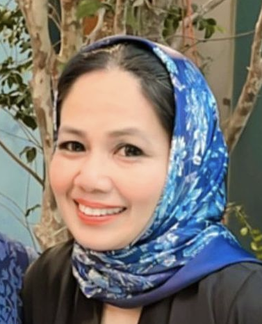 A woman with black hair wearing a blue patterned headscarf, smiling, standing outdoors with trees in the background.