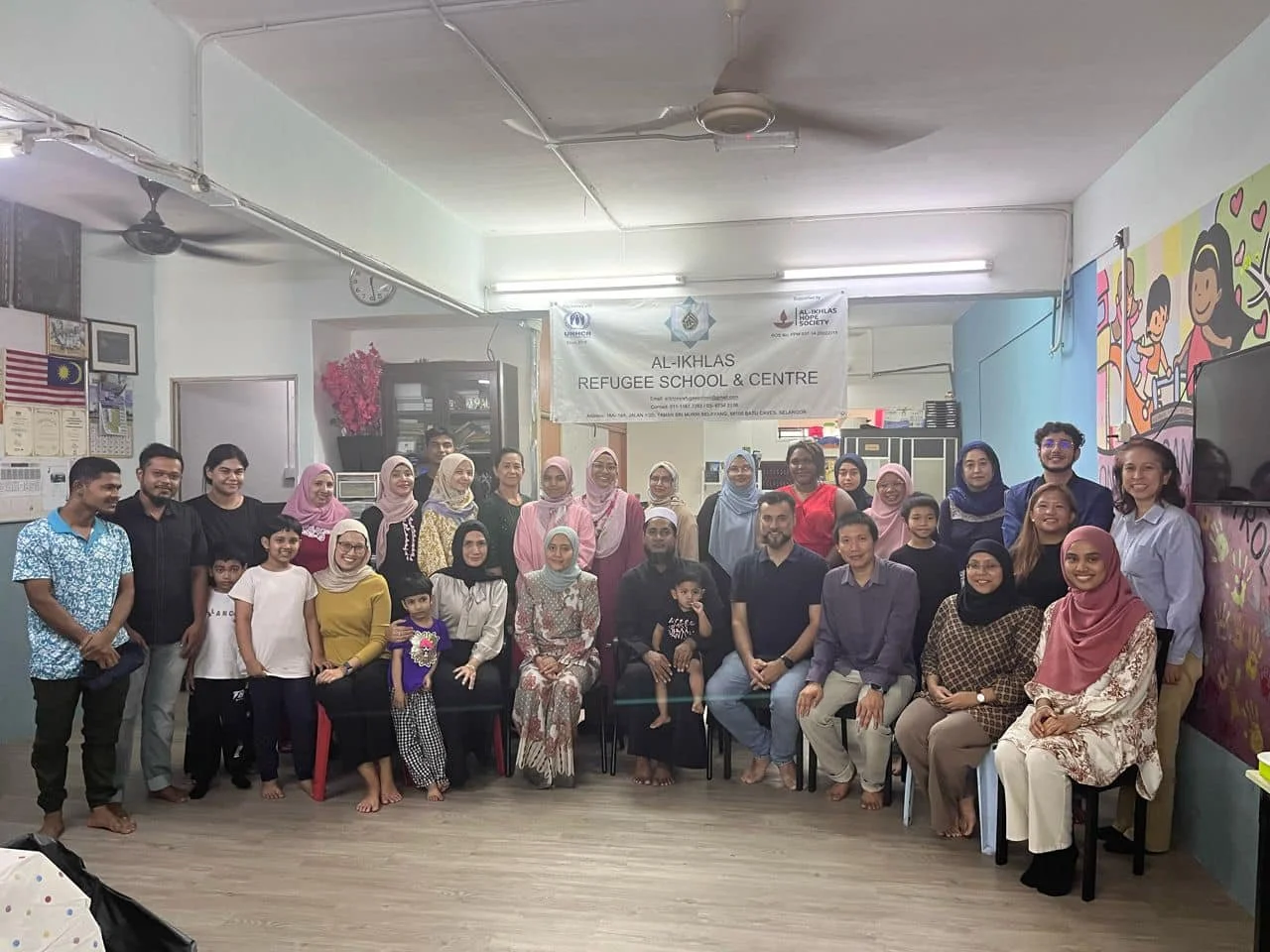 Group photo of adults and children at Al-Ikhlas Refugee School and Centre, with a banner in the background and a colorful mural on the wall.