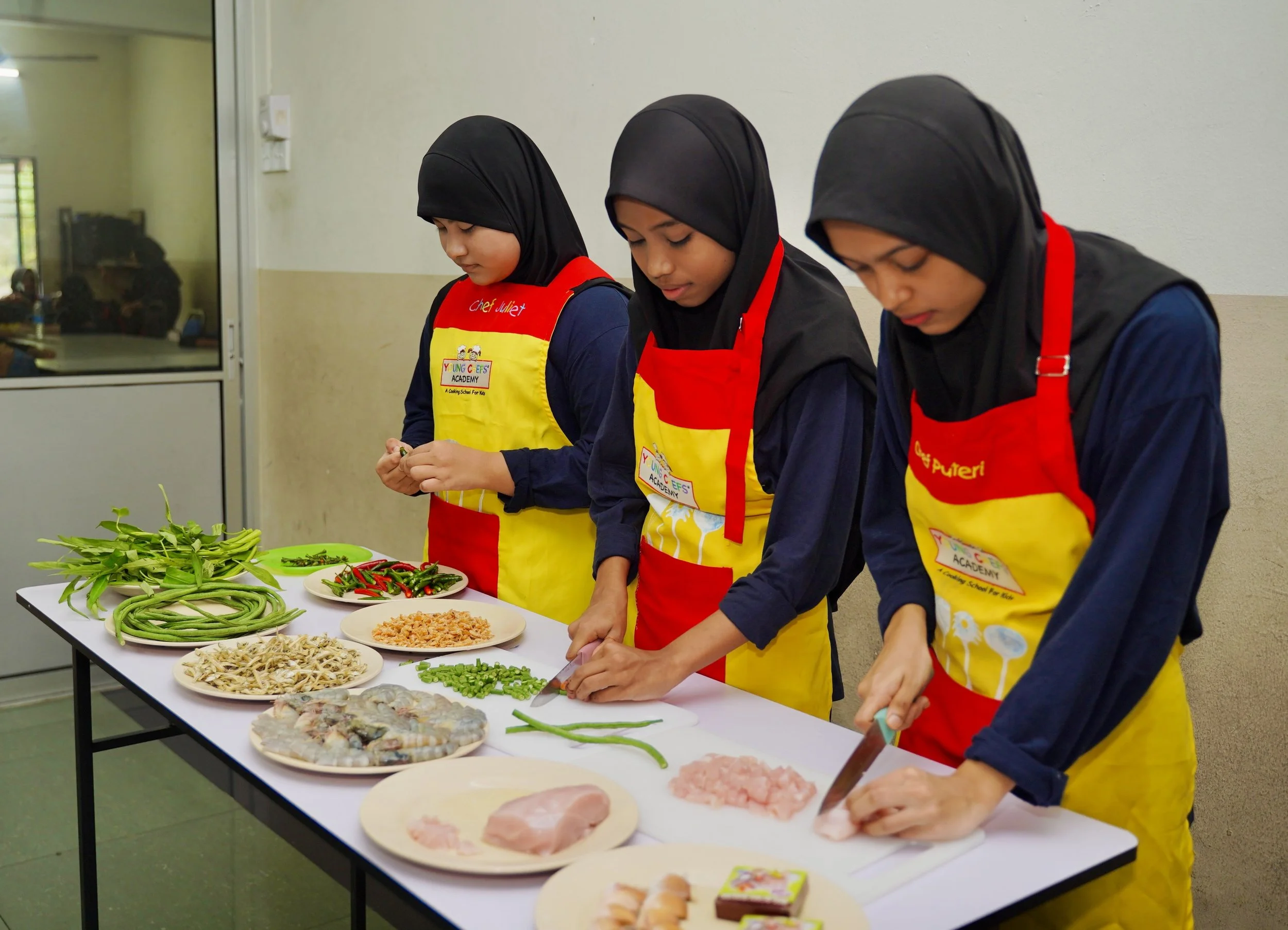 Three girls wearing black hijabs and yellow and red aprons preparing ingredients for cooking at a table. The table has green beans, red chilies, chopped fish, and chicken meat, among other ingredients.