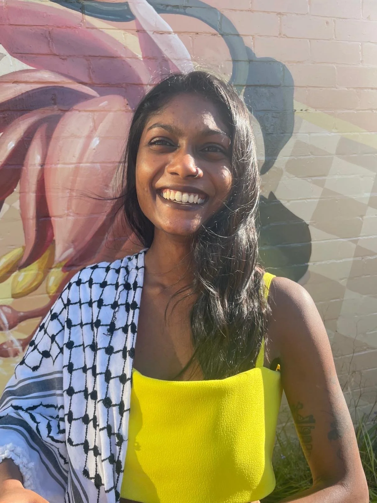 A woman with dark hair, a nose ring, and a tattoo on her arm smiling, standing in front of a colorful mural on a brick wall.
