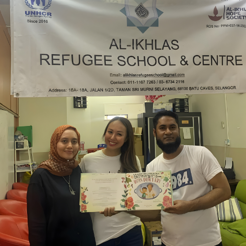 Three smiling individuals holding a colorful children's book inside a community center, with a banner for Al-Ikhlas Refugee School & Centre in the background.
