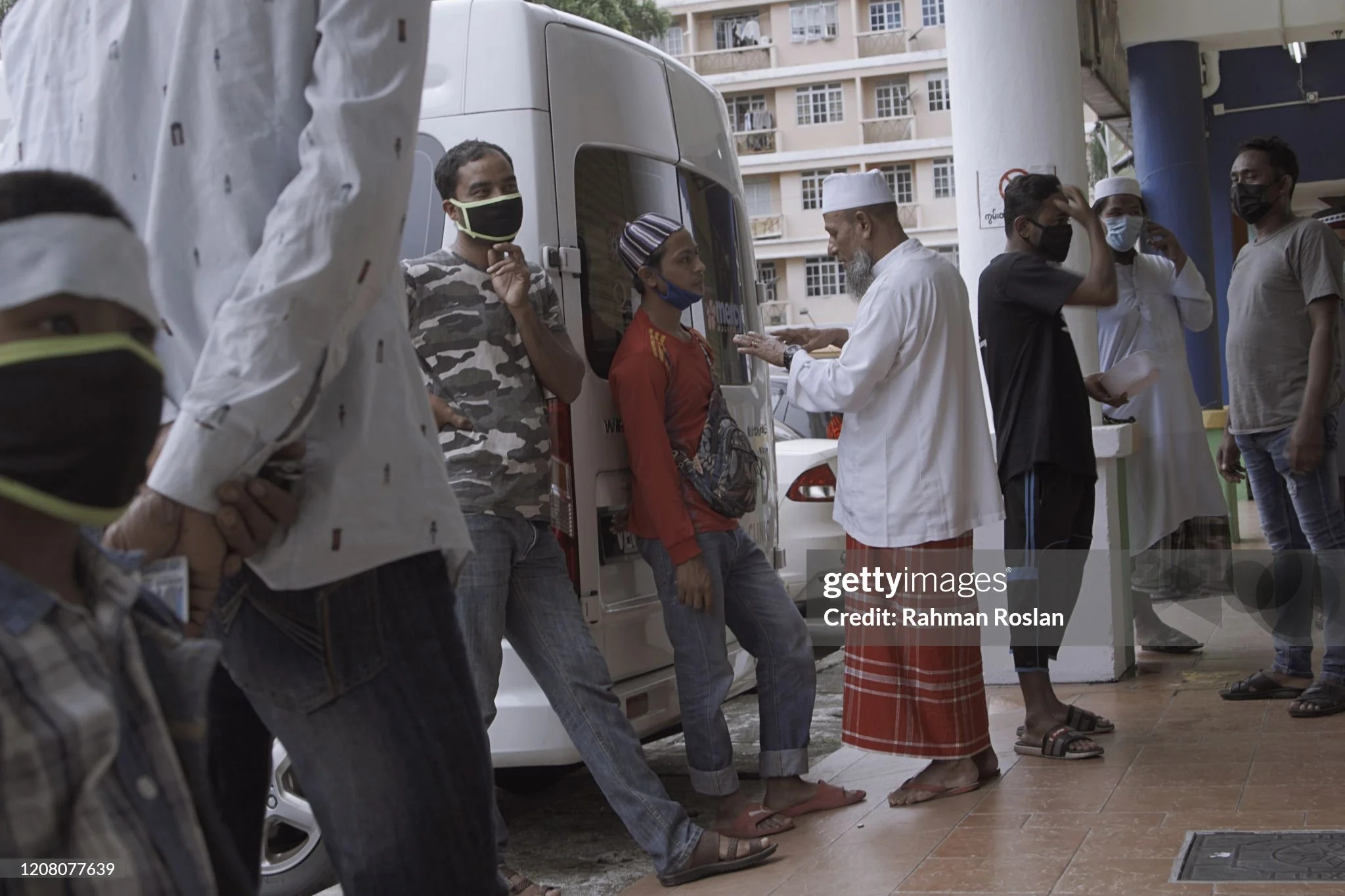 People wearing face masks standing in line outside a building, social distancing, with a man in traditional attire speaking to a woman who is leaning against a vehicle.