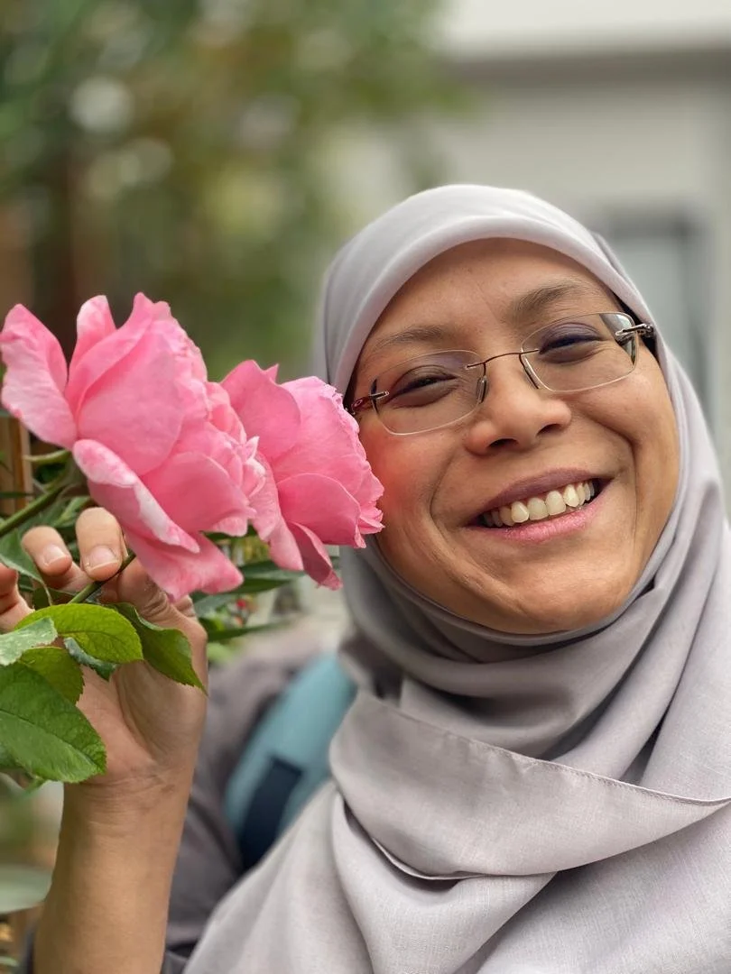 Smiling woman wearing glasses and a gray hijab holding pink flowers outdoors.