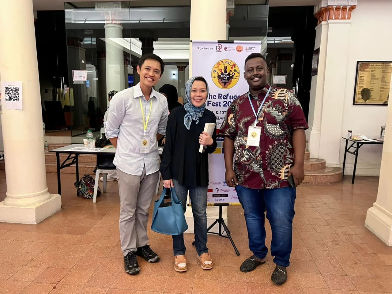 Three people standing together in front of a poster at a conference or event, smiling at the camera. The person on the left is wearing a white shirt and beige pants, the individual in the middle is in a black jacket and headscarf, holding a blue handbag, and the person on the right is dressed in a patterned maroon shirt and blue jeans.