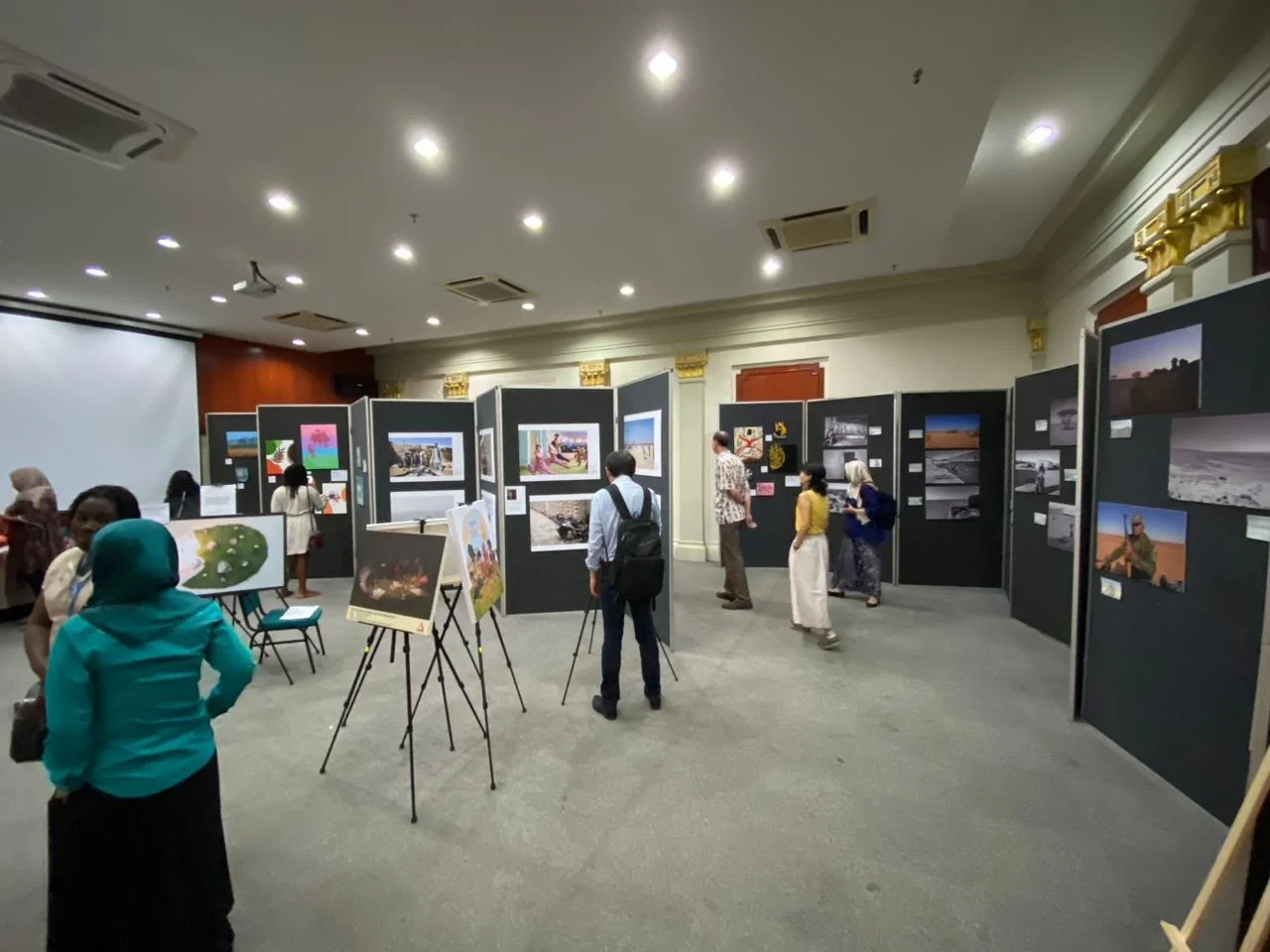 People viewing photographs and artwork displayed on black foldable display panels in an indoor exhibition hall.