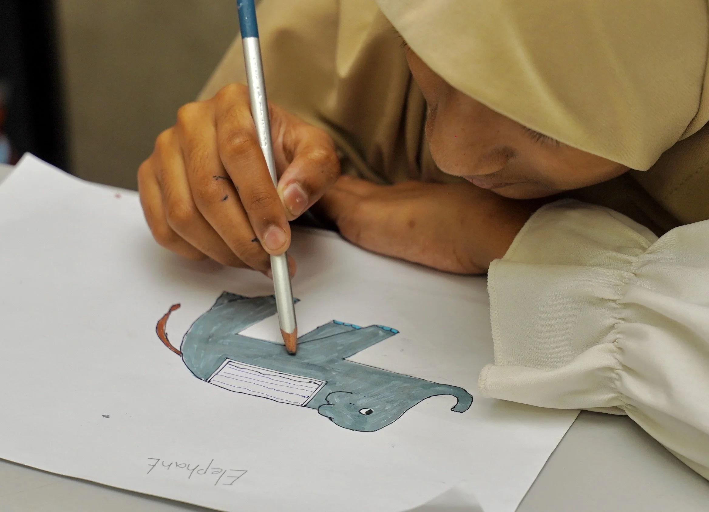 A person wearing a beige hijab is drawing a colorful elephant on a white sheet of paper, using colored pencils, with their left arm resting on the table.