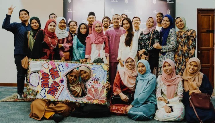 Group of diverse young adults, mostly women, smiling and posing together indoors at a celebration or event, with two women holding a colorful handmade sign with a heart, notebook, and creative decorations.