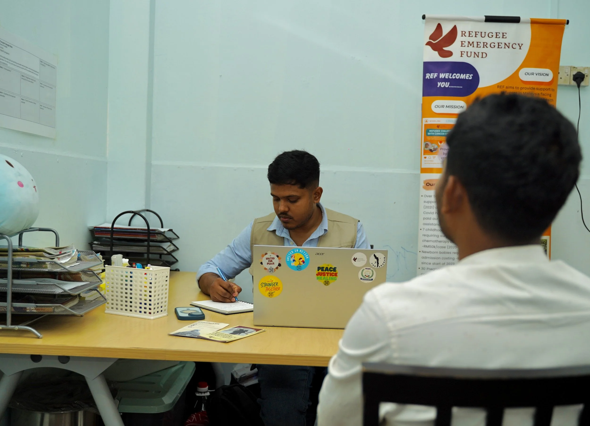 Two men in an office. One is sitting at a desk with a laptop decorated with stickers, writing in a notebook. The other person, seen from behind, is wearing a white shirt and is facing the person at the desk. The office has a poster on the wall with the title 'Refugee Emergency Fund' and a globe on the desk.