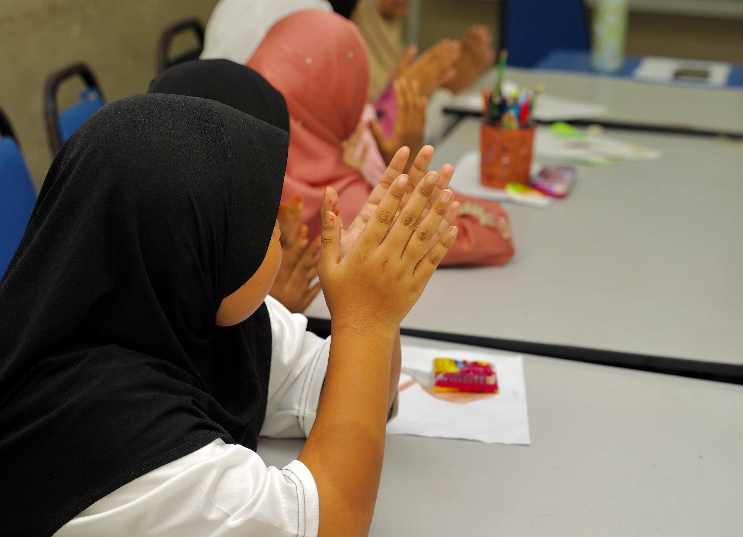 Group of children sitting at a table with hands raised, participating in a classroom activity. Some children are wearing headscarves.