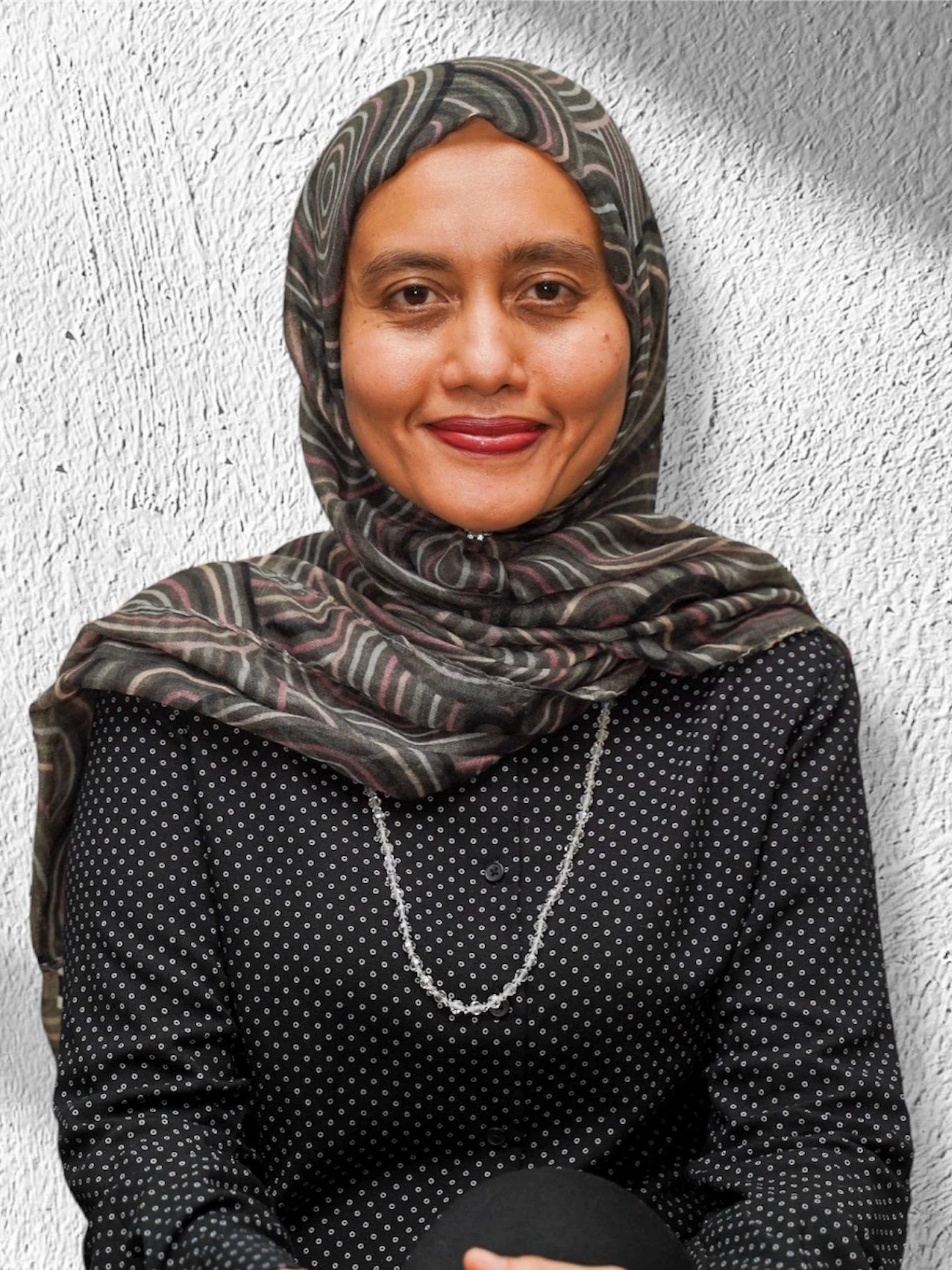 A woman wearing a patterned hijab and black polka dot shirt, smiling, against a textured white wall.