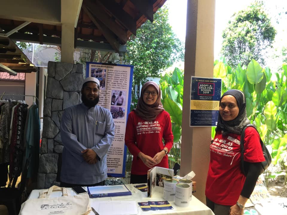 Three people standing behind a table at an outdoor event, with posters and informational materials on the table and large green plants in the background.