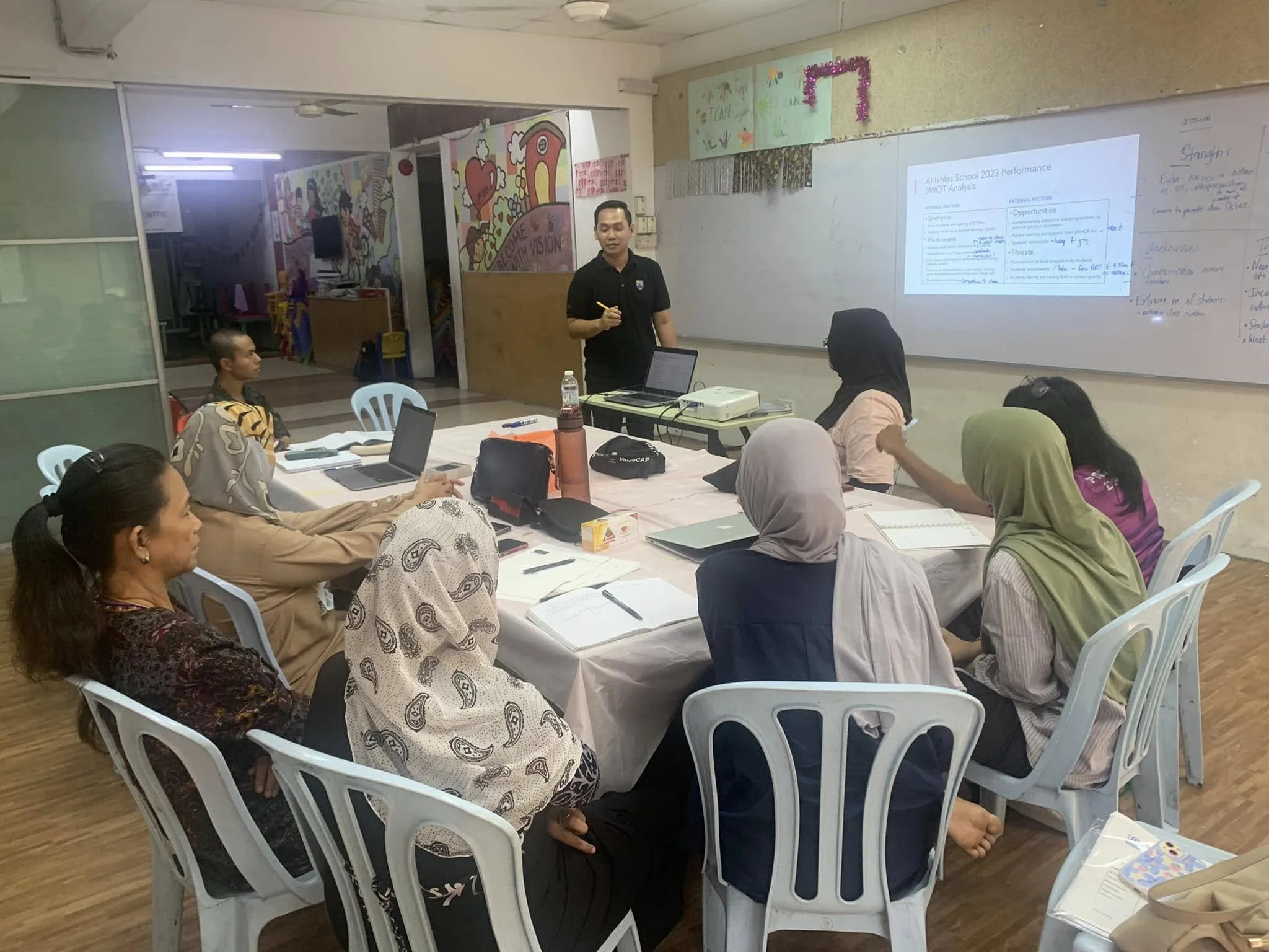 A group of women seated around a table in a classroom, attending a presentation led by a man standing in front of a whiteboard with a slide projected on it.
