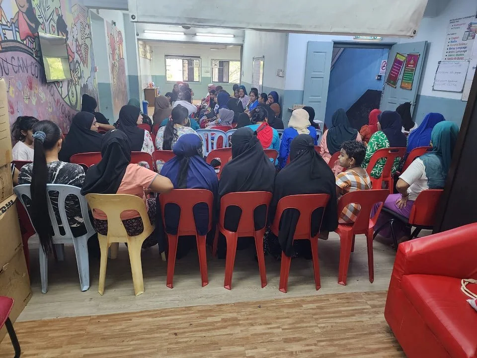 A group of women and children attending a meeting or class in a room with colorful wall murals, plastic chairs, and a door leading outside.