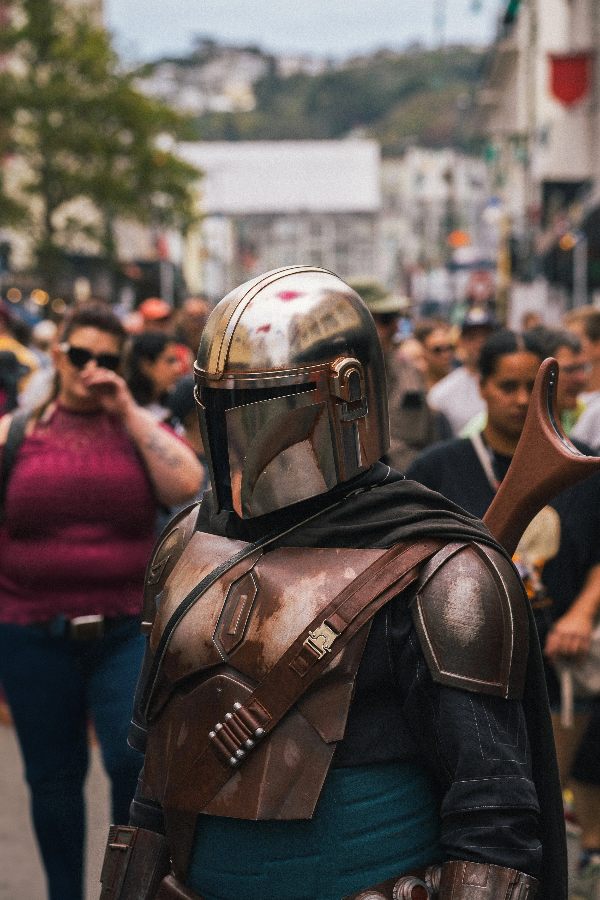 A person dressed as Boba Fett from Star Wars, wearing a metallic helmet and armor, standing among a crowd in an outdoor city setting.
