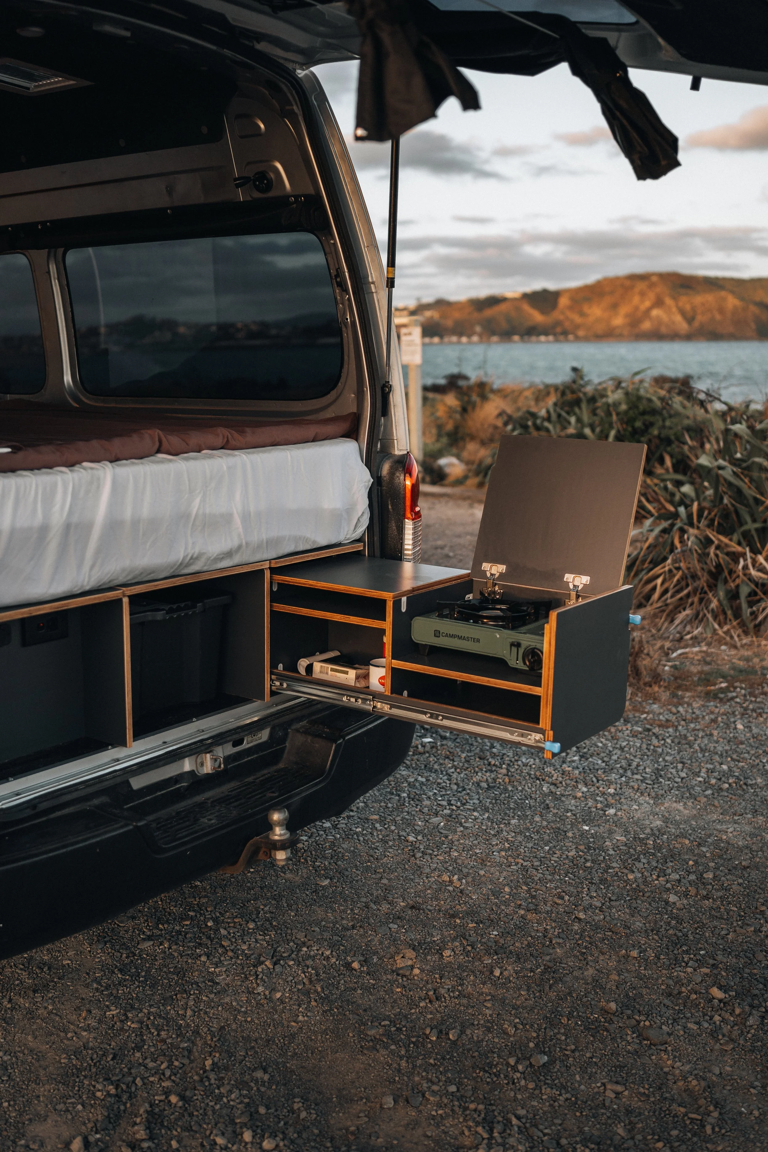 Open van with built-in wooden kitchen and portable stove, parked near a scenic lakeside or coastal area at sunset.
