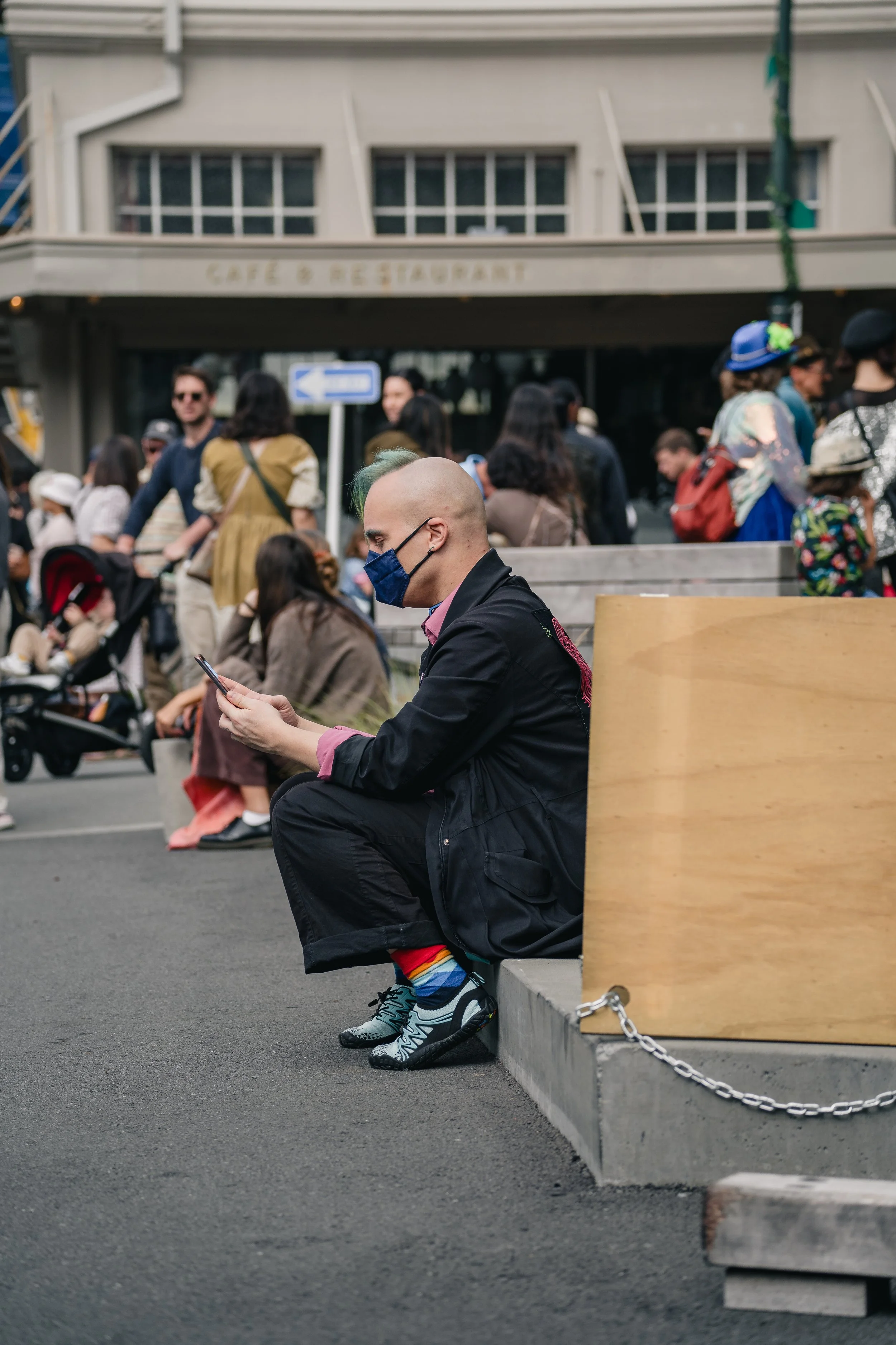 A man with a shaved head, wearing a blue face mask, black jacket, colorful rainbow socks, sneakers, and glasses, sitting on a gray concrete bench in a busy urban area, looking at his phone. Behind him, a crowd of people including children and adults,