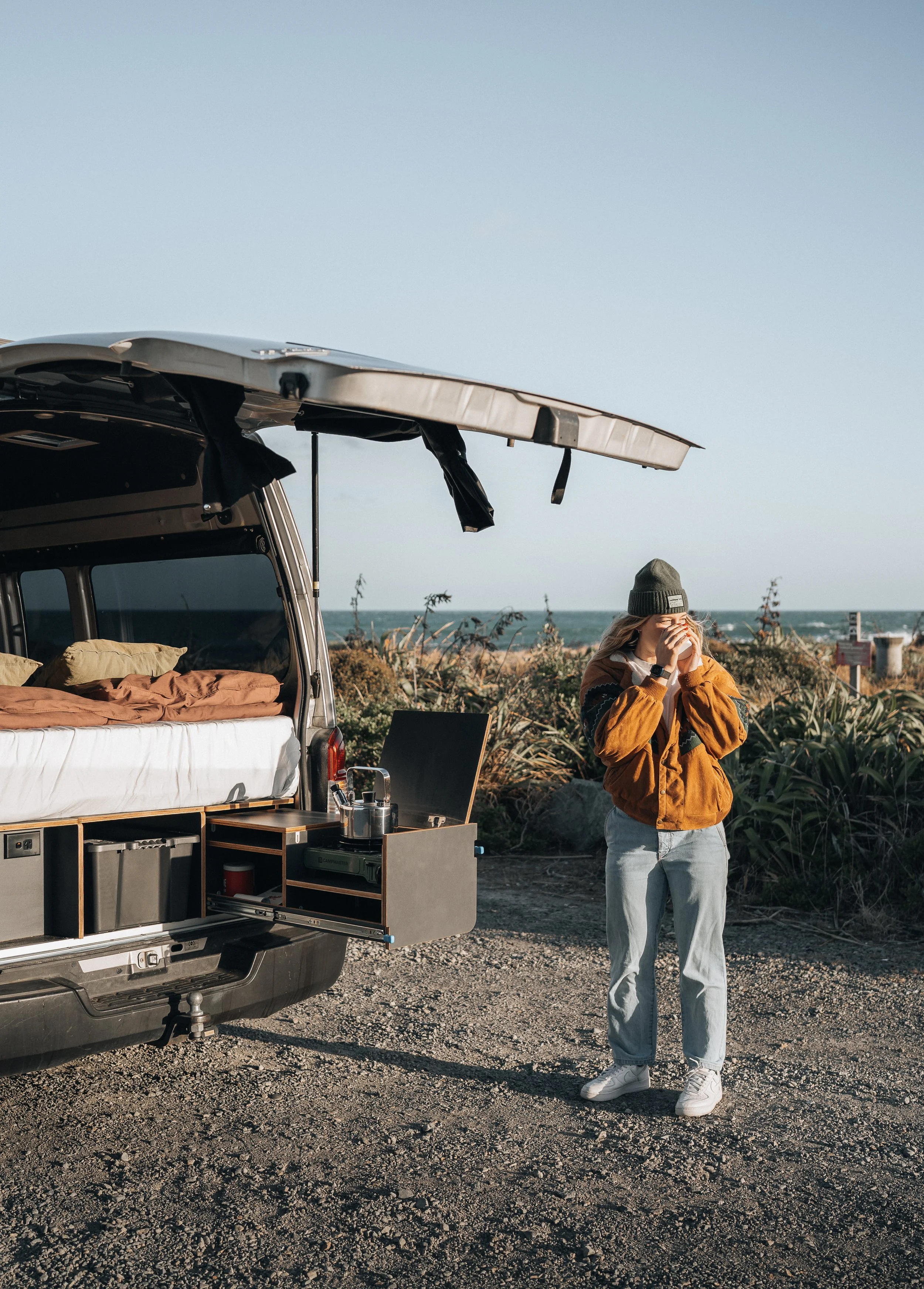 A woman standing next to an open camper van at a beach, holding a cup and wearing a beanie and casual clothing.