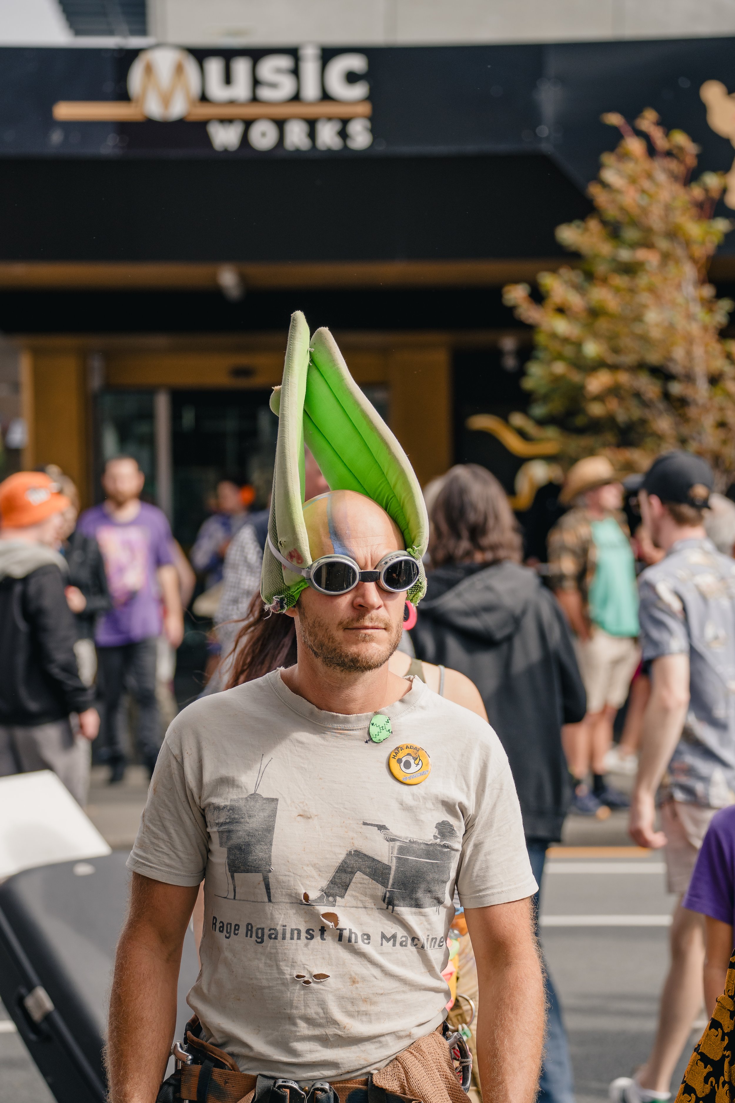 A man wearing goggles and a large, green, leaf-shaped hat at a crowded outdoor event.