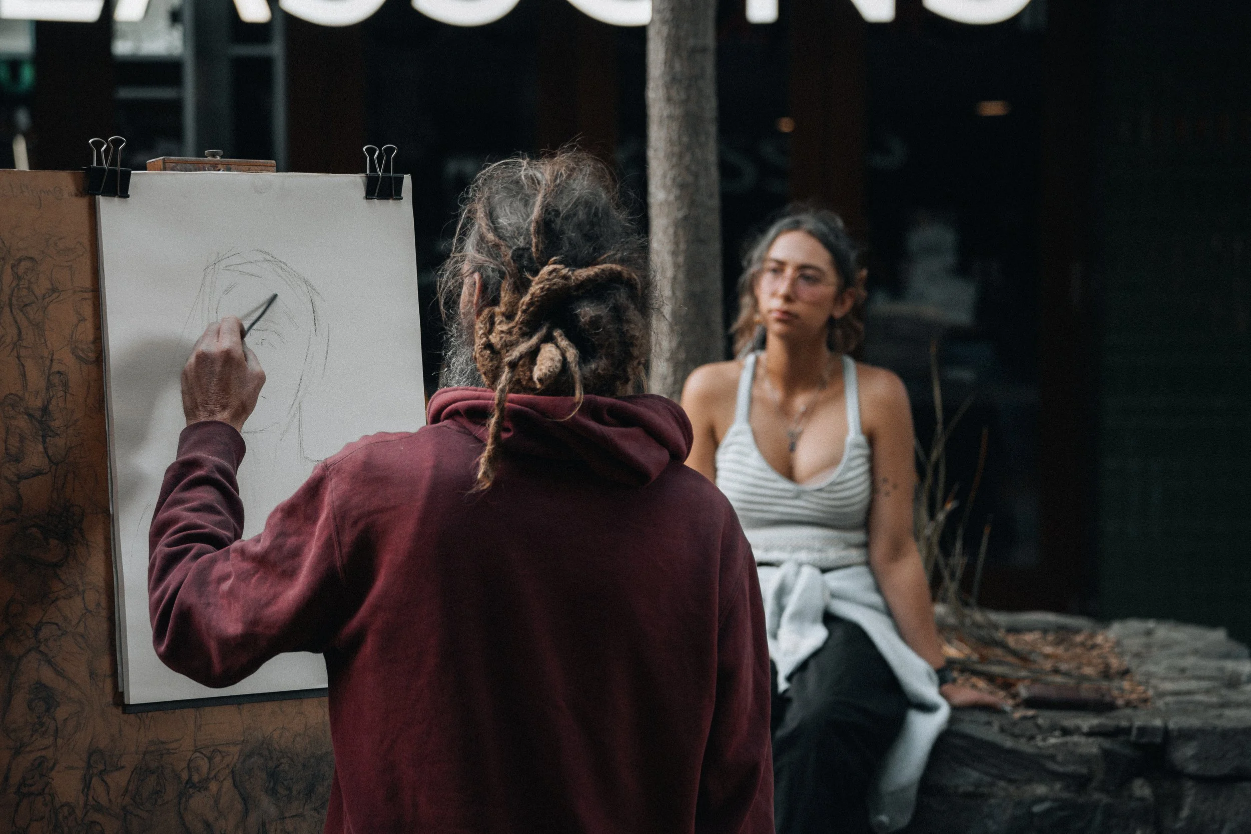 An artist with dreadlocks is sketching a portrait on a white canvas in an outdoor setting, while a woman with glasses and tattoos sits on a stone ledge in the background.