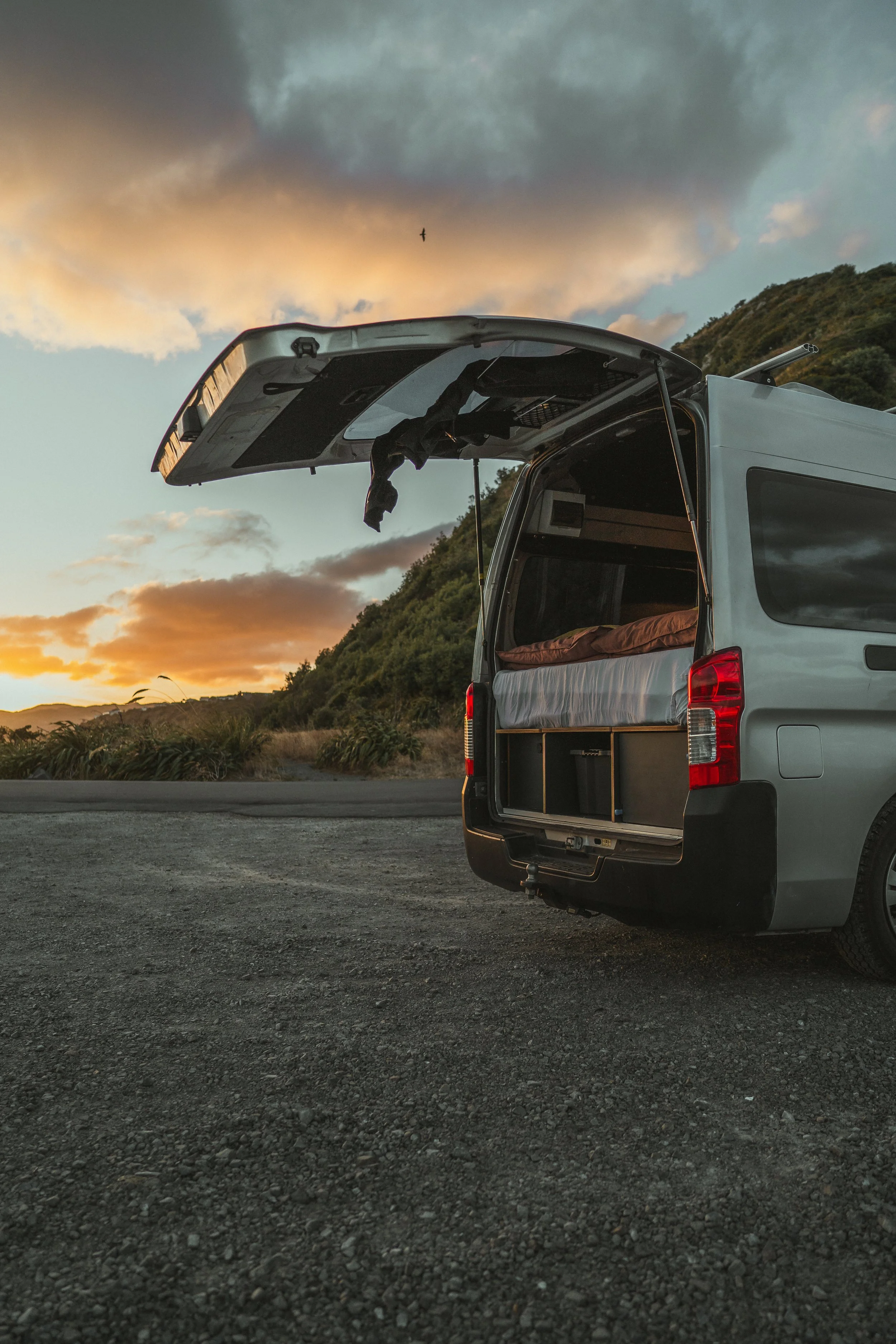Rear view of a camper van parked on a gravel road at sunset, with the rear door open revealing a bed inside and a landscape with hills and a partly cloudy sky.
