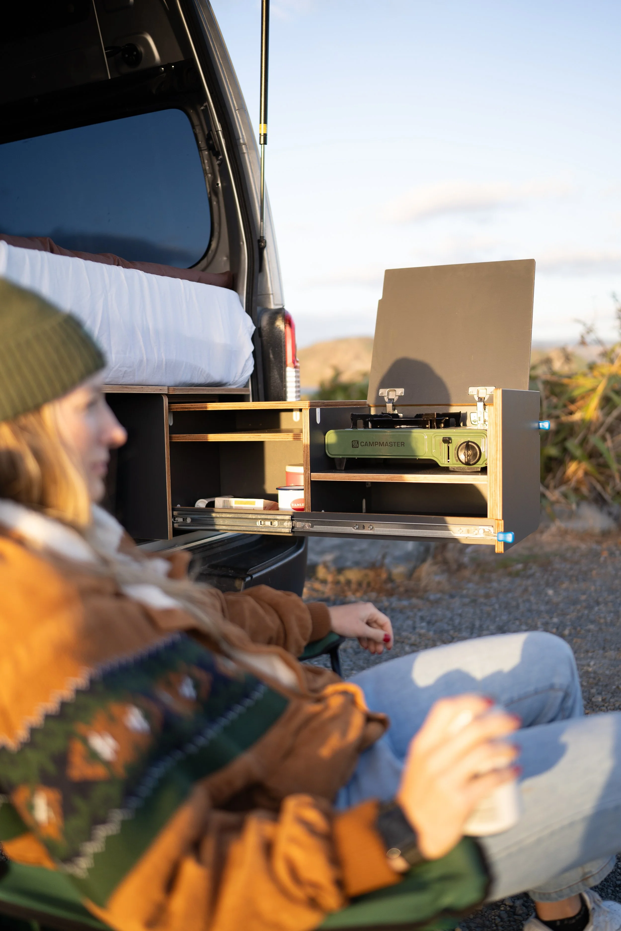 A woman sitting on a camping chair near the open back of a camper van, with a portable stove and camping supplies inside the vehicle.