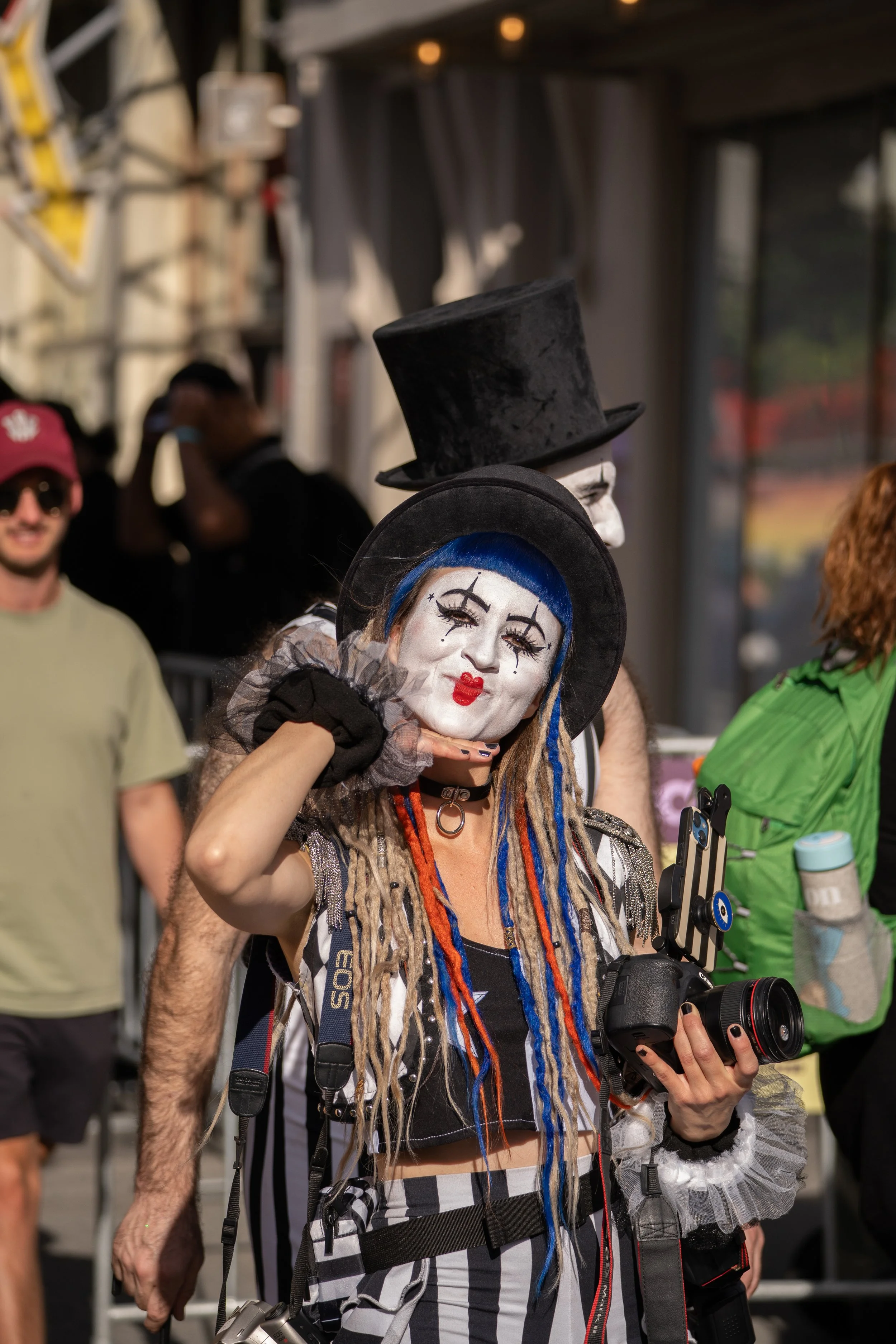 A person dressed as a clown with face paint, blue dreadlocks, and striped pants, holding a camera and posing with a person in a mime costume wearing a tall black hat and white face paint at an outdoor event.